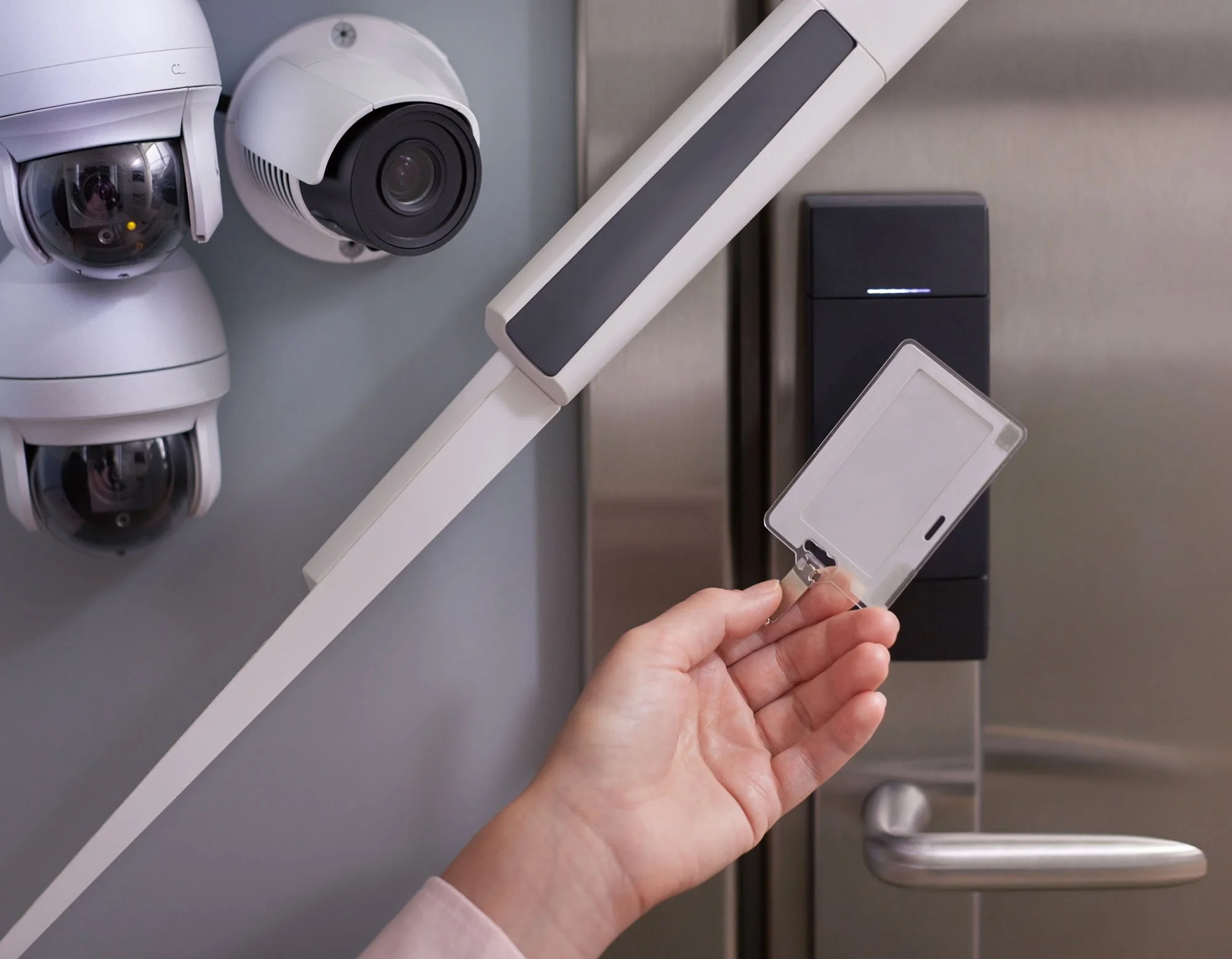 A person holding a transparent keycard near a door lock. On the wall are three security cameras and a doorbell, with a modern metallic door in the background.