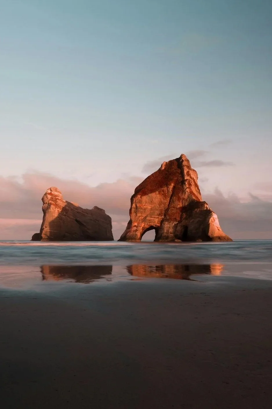 Two large rock formations in the ocean near the shore during sunset, with reflections on the wet sand.