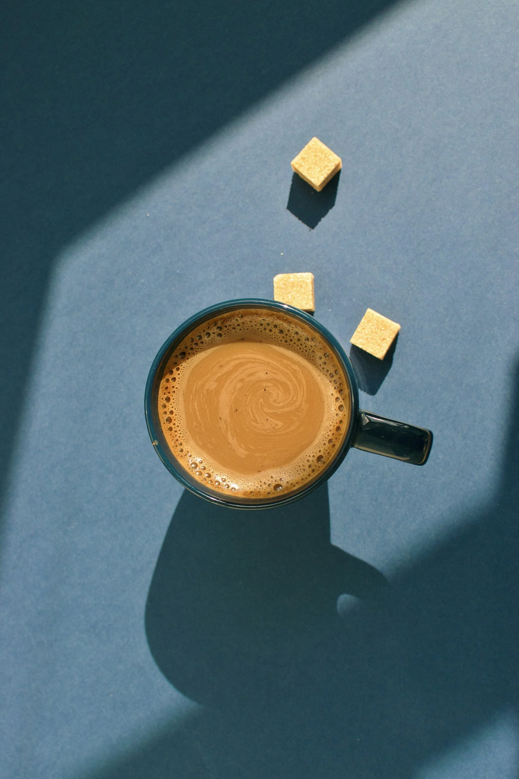 A top-down view of a black coffee mug filled with coffee on a blue surface, with three sugar cubes nearby and shadow cast from the mug.