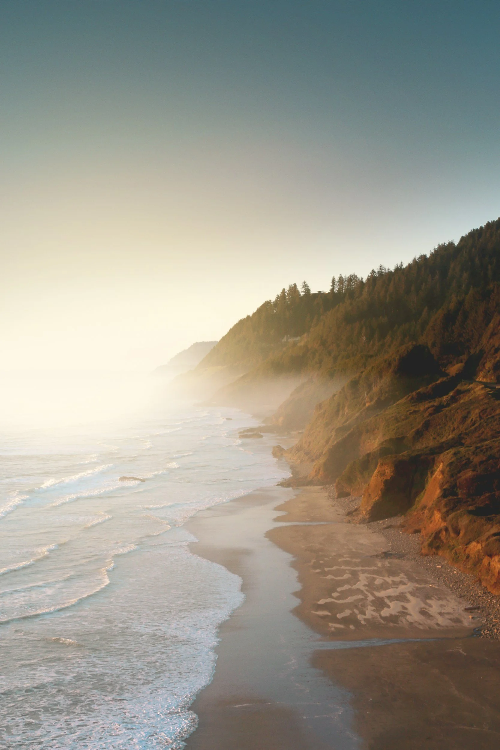 Sunset or sunrise over a beach with waves, sandy shore, and rocky cliffs with trees.