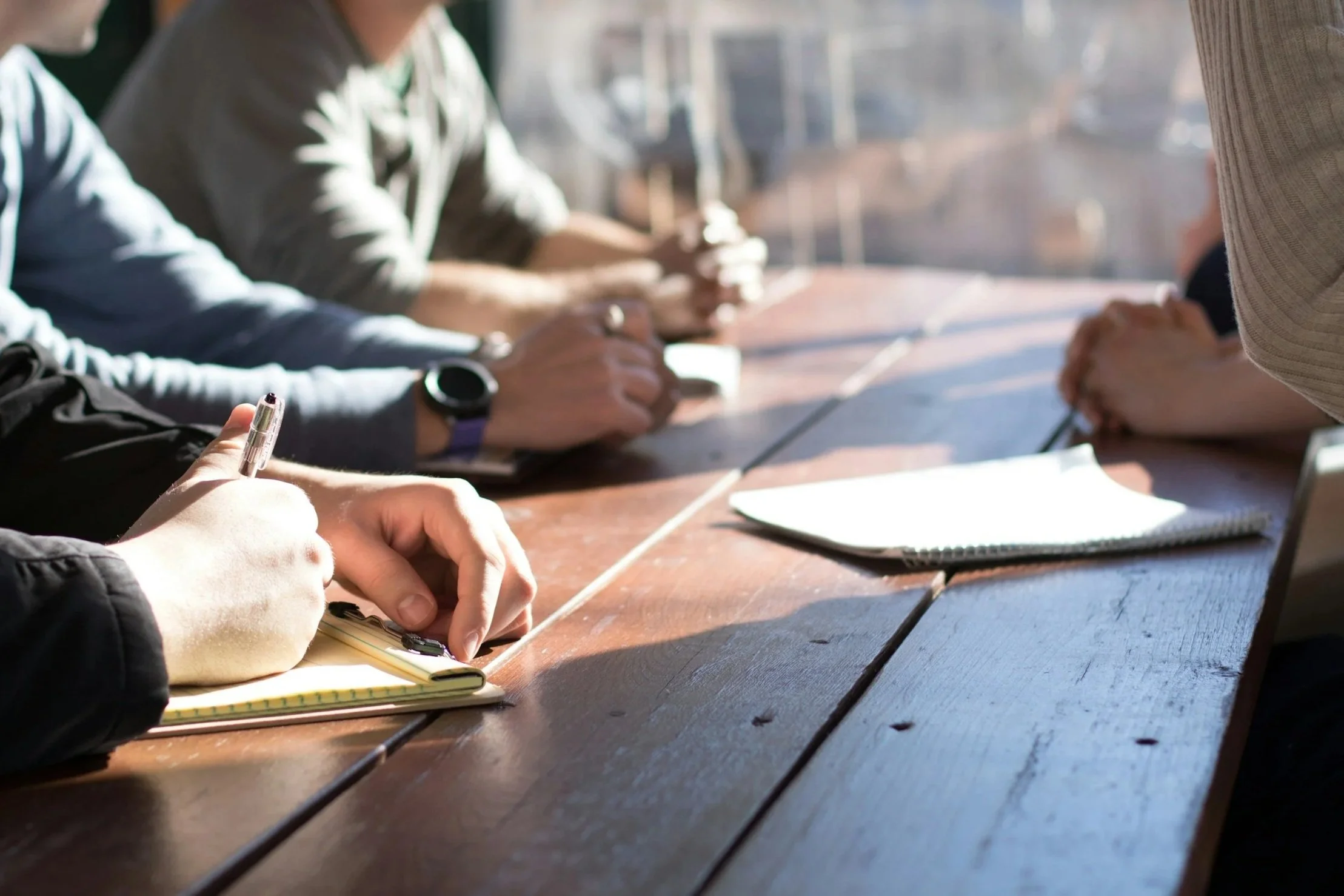 People sitting at a wooden table with notebooks, some writing, in a well-lit room.