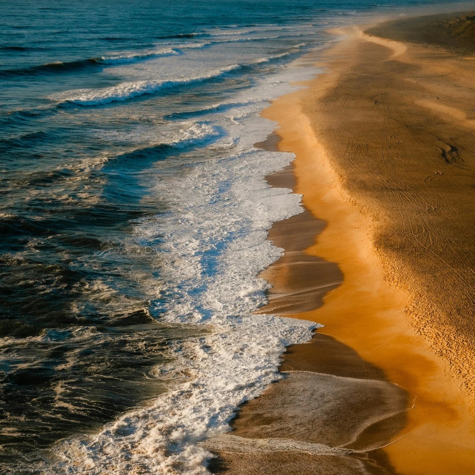 An aerial view of a sandy beach with ocean waves crashing onto the shore during sunset.