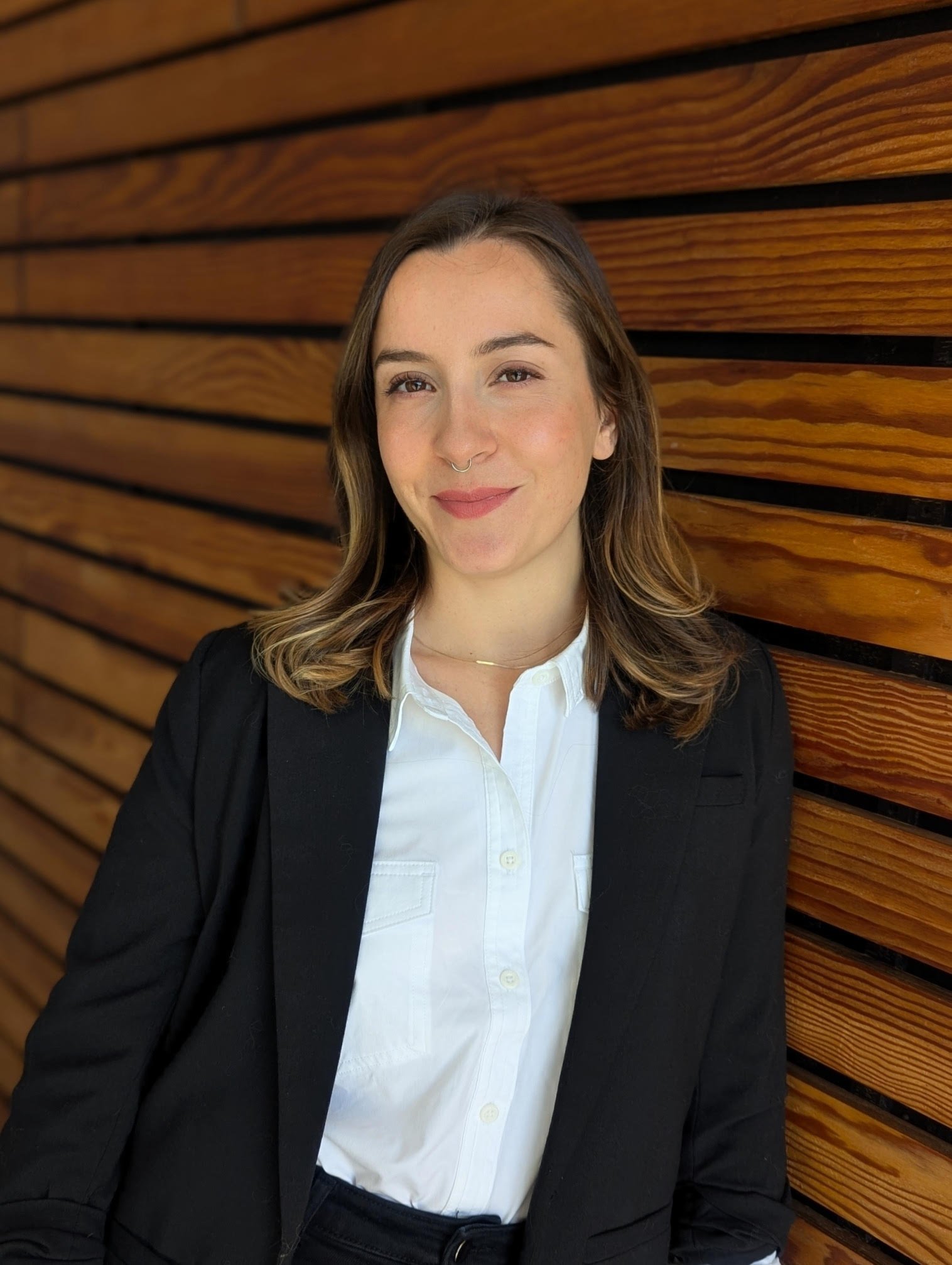 A woman with shoulder-length brown hair and a septum piercing standing against a wooden wall, wearing a white shirt and black blazer, smiling slightly at the camera.