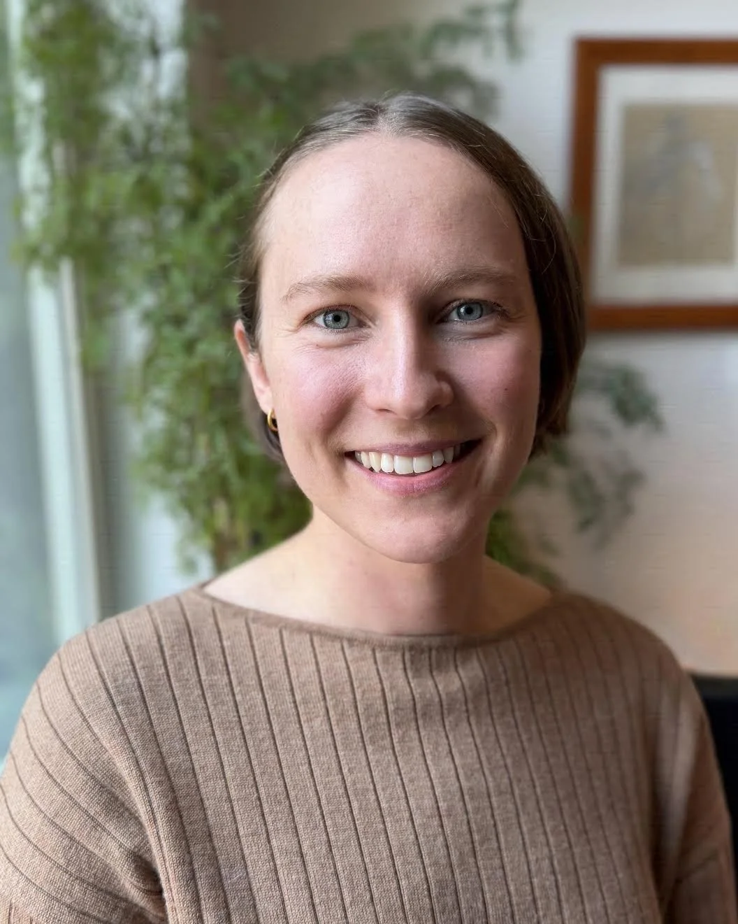 A woman with short brown hair, blue eyes, and a bright smile, wearing a tan ribbed top, standing indoors near a window with plants and framed art in the background.