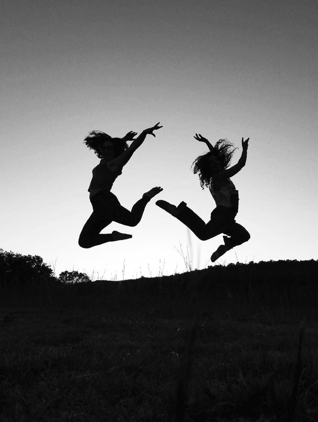 Two people jumping in the air, silhouetted against a sunset sky, with grass and hills in the background.