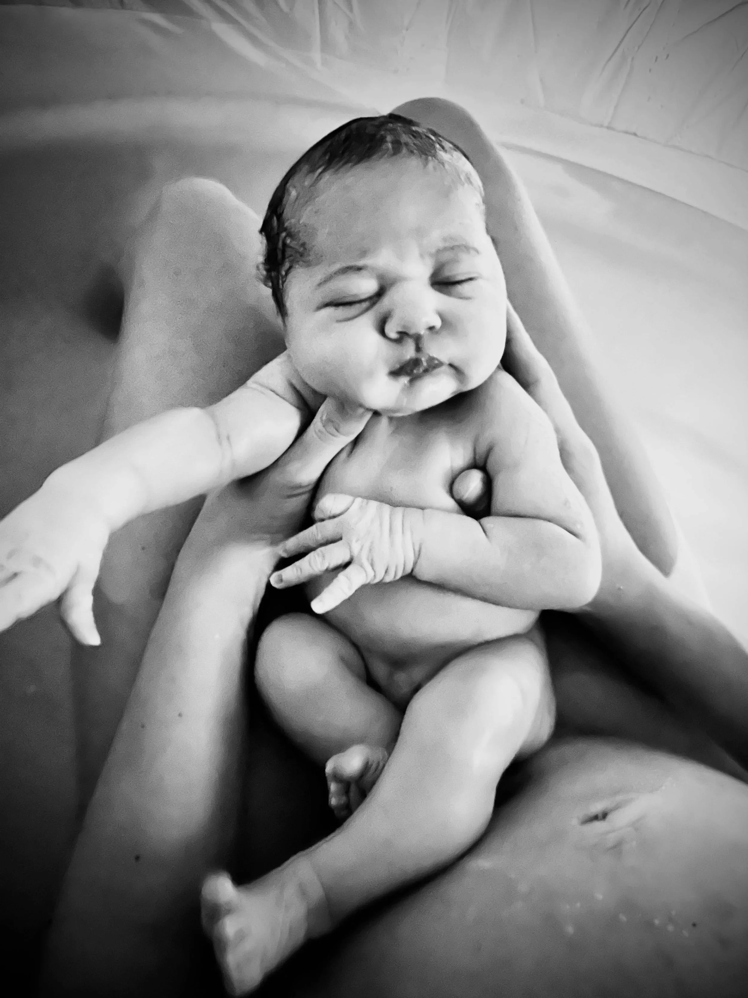 Black and white photo of a newborn baby being held by an adult, with the baby's eyes closed and a peaceful expression, in a bath or similar setting.