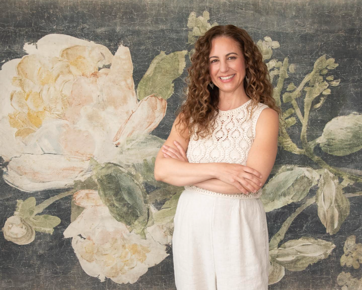A woman with curly brown hair wearing a sleeveless lace white top and light-colored skirt, standing with arms crossed and smiling in front of a floral mural background.