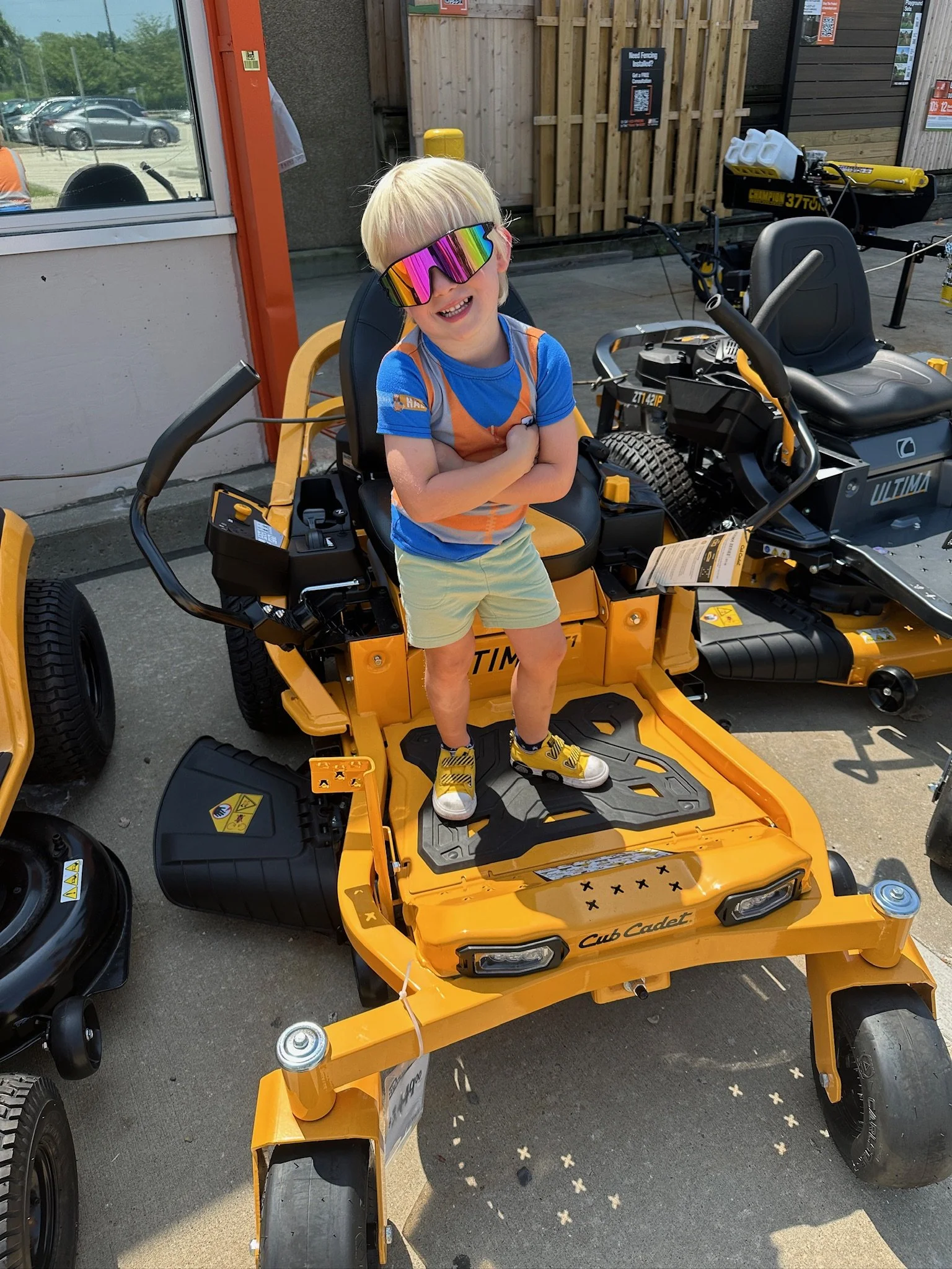 A young boy with blonde hair and wearing rainbow sunglasses, a blue T-shirt, and yellow shorts, smiling and standing on a yellow Cub Cadet ride-on lawn mower in an outdoor store parking lot.
