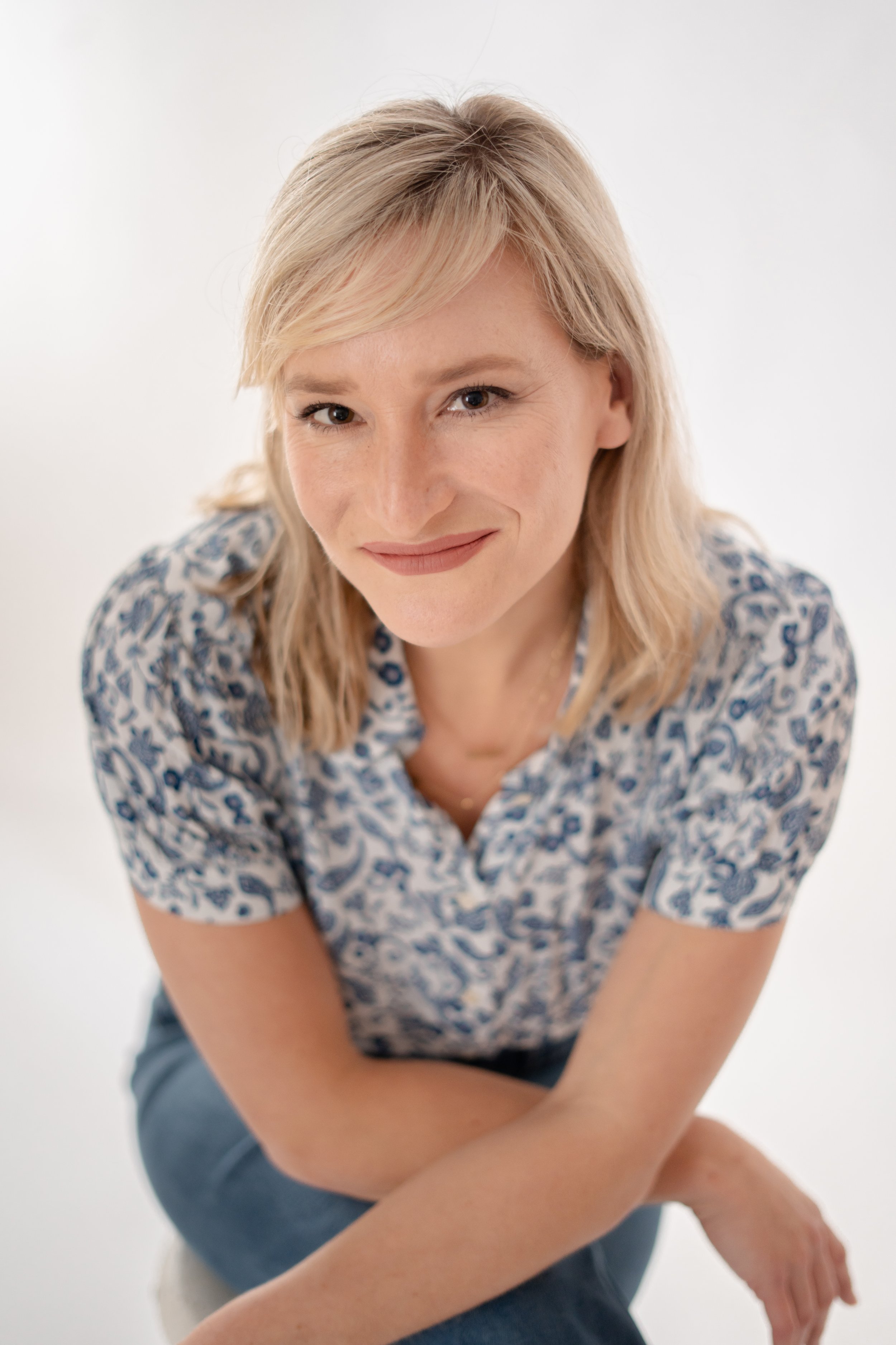 A woman with blonde hair and fair skin, smiling and looking directly at the camera, wearing a patterned blouse and seated against a plain white background.