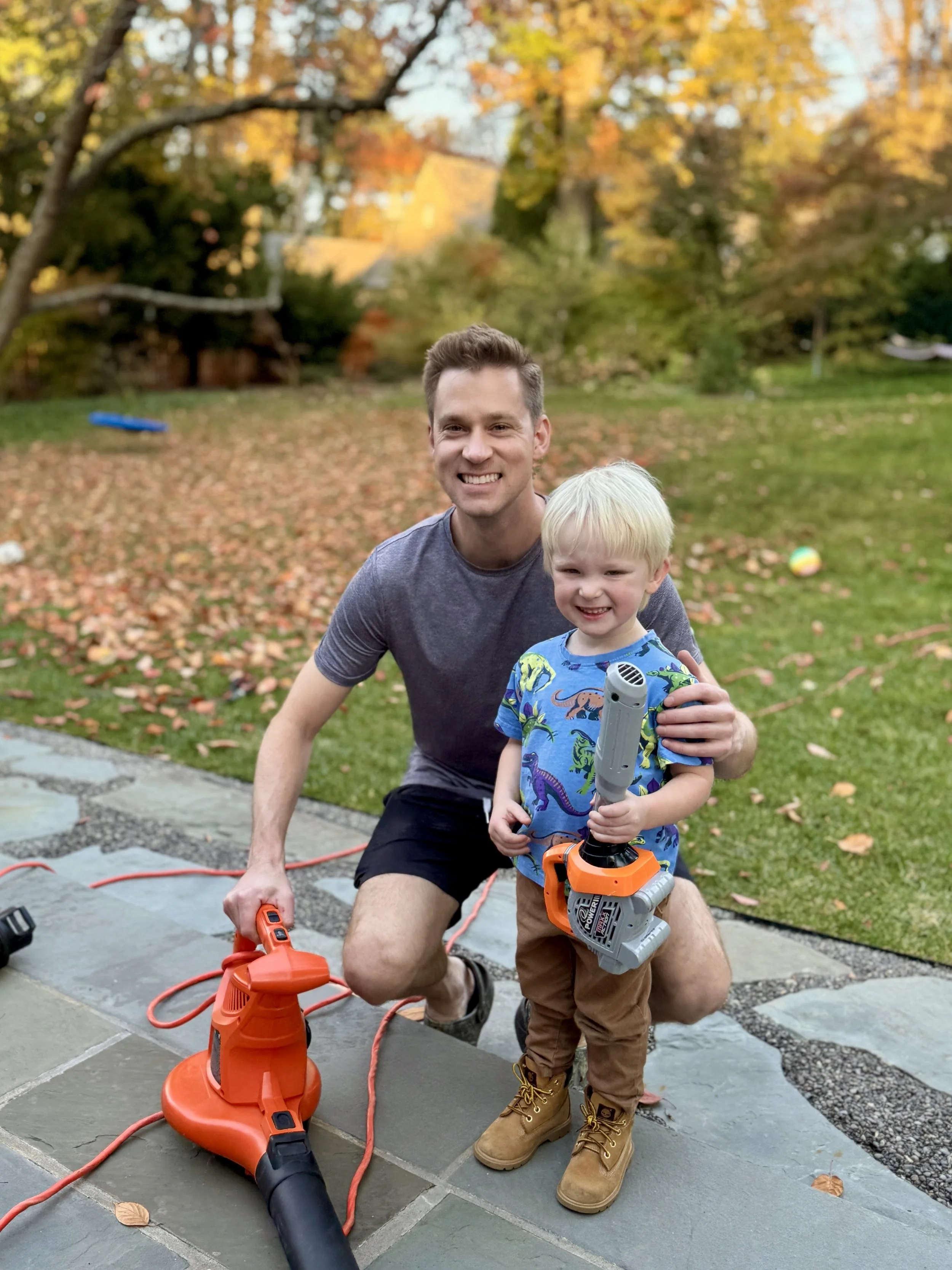 A man and a young boy smiling outdoors on a fall day. The boy is holding a toy chainsaw and the man is holding a leaf blower. There are fallen leaves on the ground and trees with autumn foliage in the background.