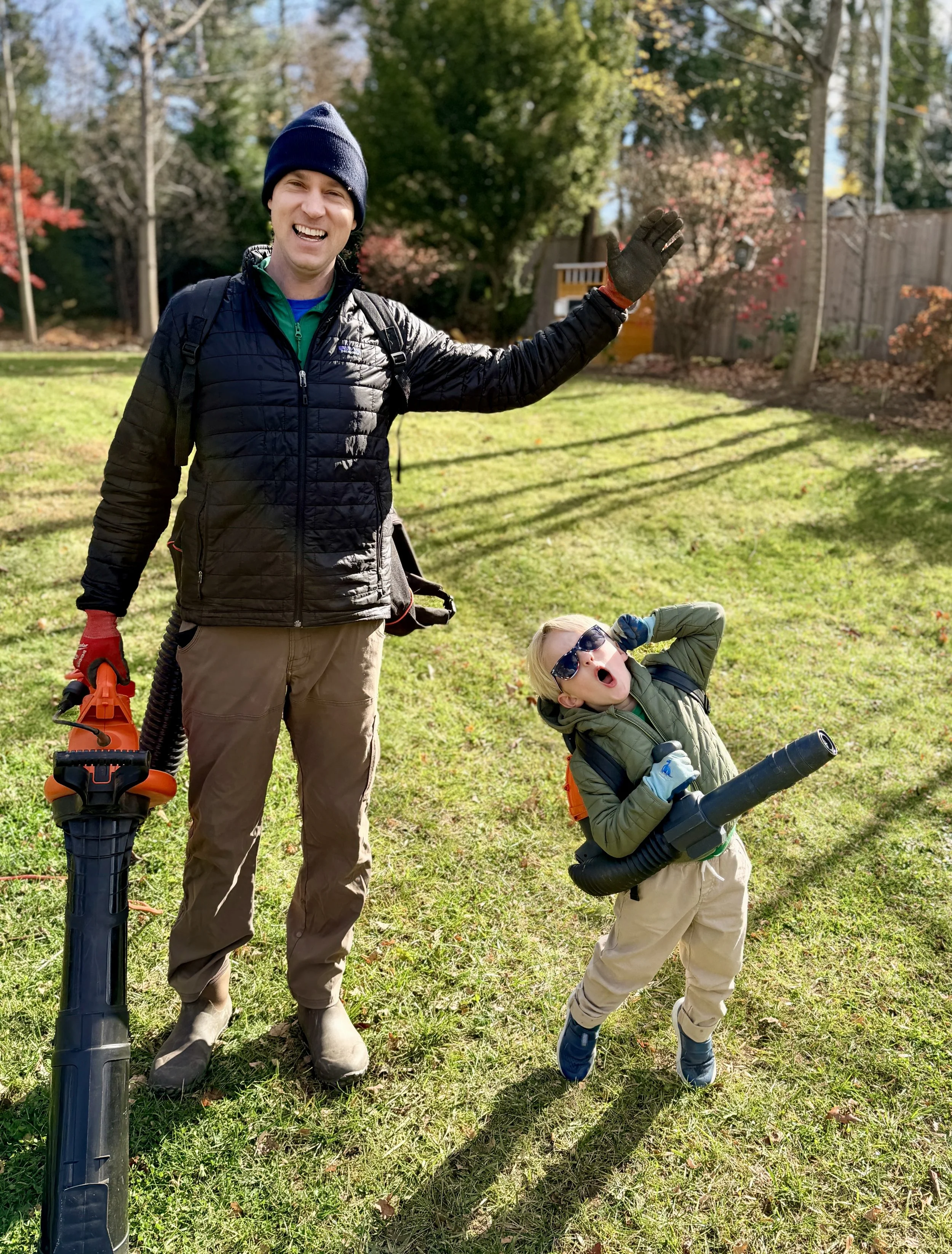 A man and a boy standing outdoors on grass, both smiling and posing with gardening equipment, with trees and a fence in the background on a sunny day.