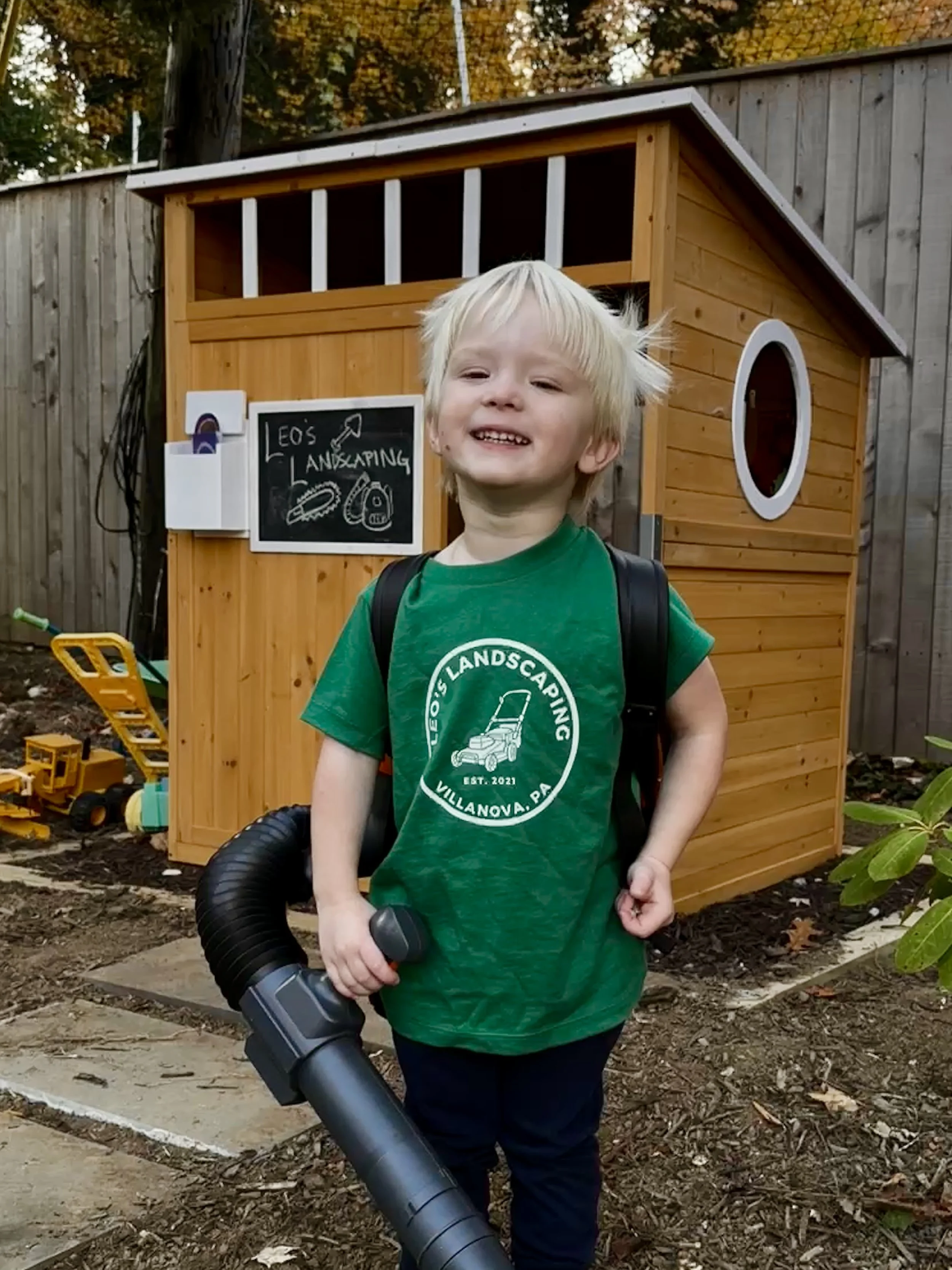 A young boy with blond hair smiling and holding a leaf blower in a yard with a playhouse in the background. The playhouse is made of wood with a small circular window and a chalkboard sign that reads "Leo's Landscaping" and features drawings of a shovel, gloves, and a lawnmower. There are toys and gardening tools scattered around.
