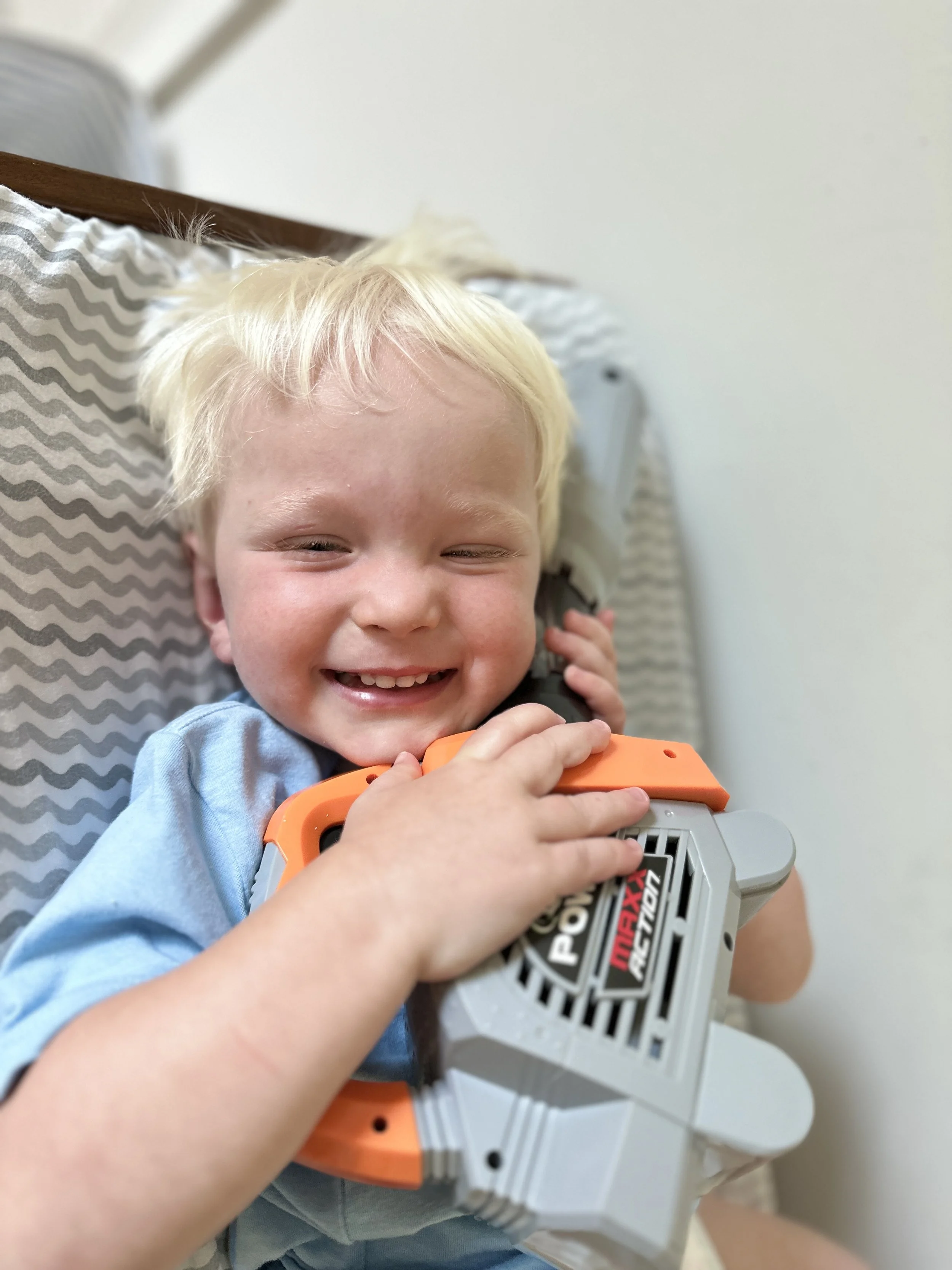 A smiling young boy with blonde hair lying on a bed, hugging a Nerf blaster toy.