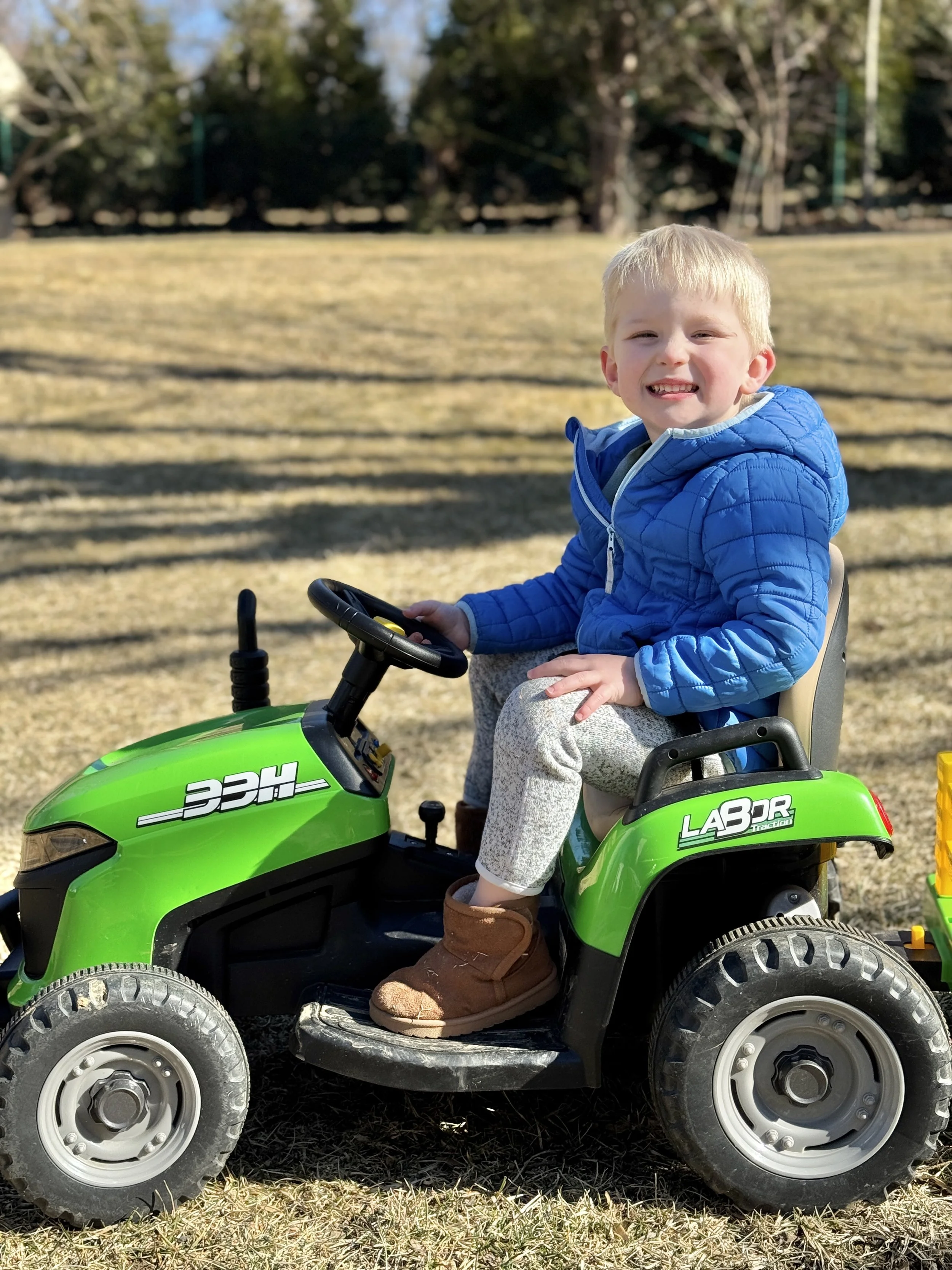 A young boy with blonde hair smiling while sitting on a green toy car outdoors, wearing a blue jacket and brown boots, with a background of trees and a grassy field.