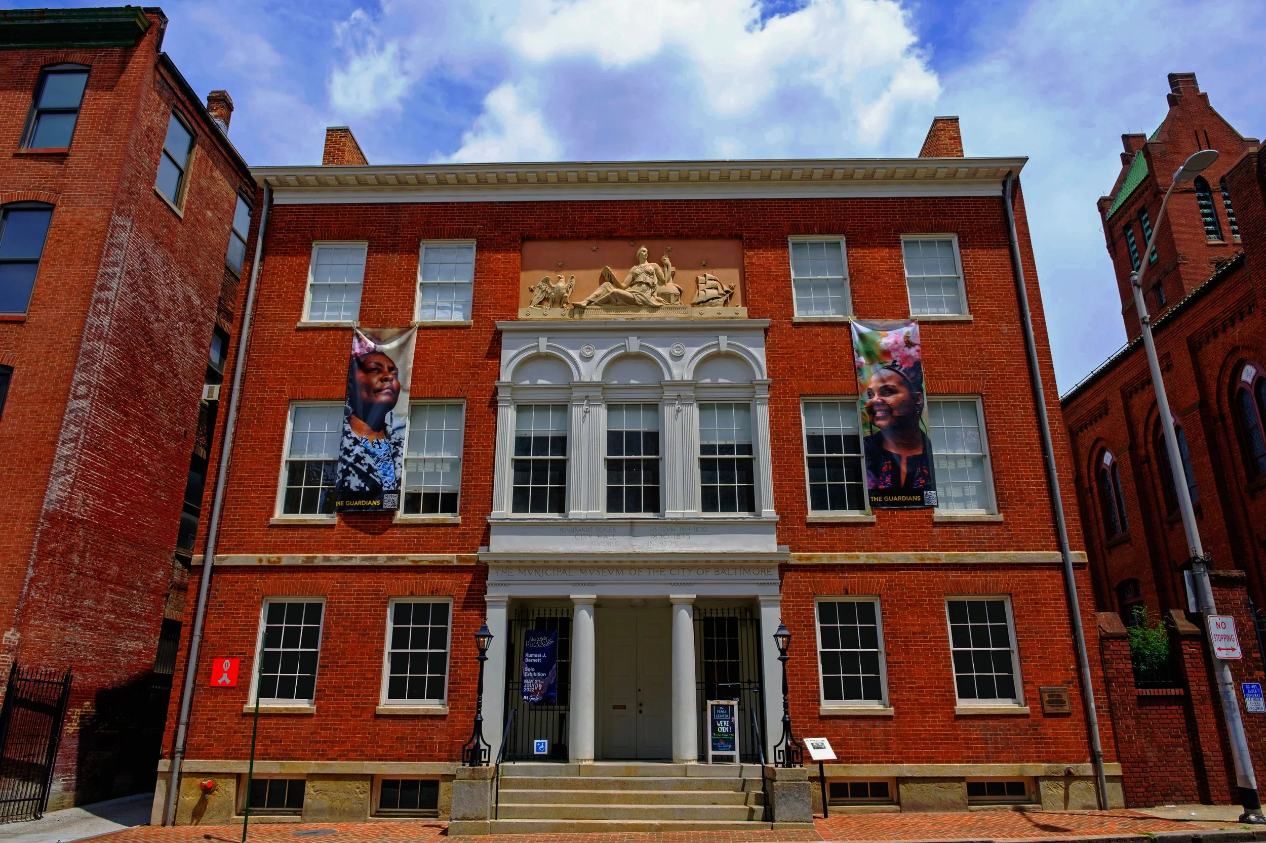 Front view of the Baltimore Municipal Museum, a red brick building with classical columns and sculptures above the entrance, two banners on either side of the windows, and a streetlamp on the right.