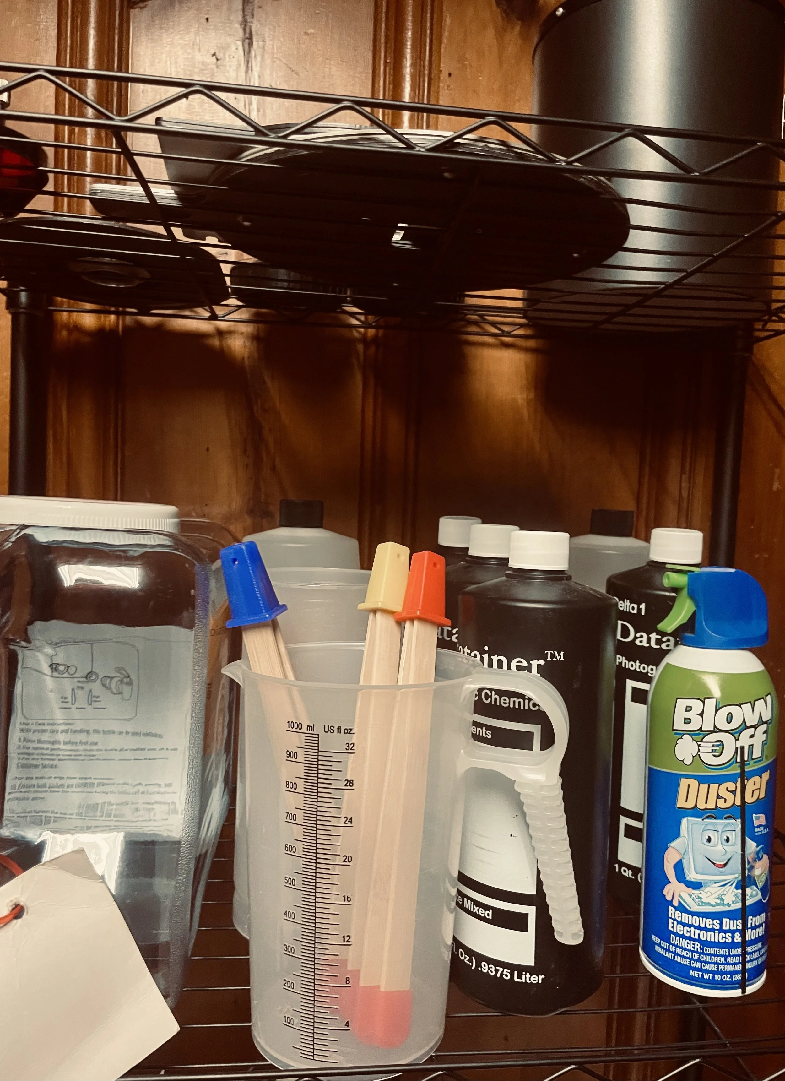 Laboratory supplies on a metal shelf, including a beaker with colored sticks, bottles of chemicals, and a can of dust cleaner.