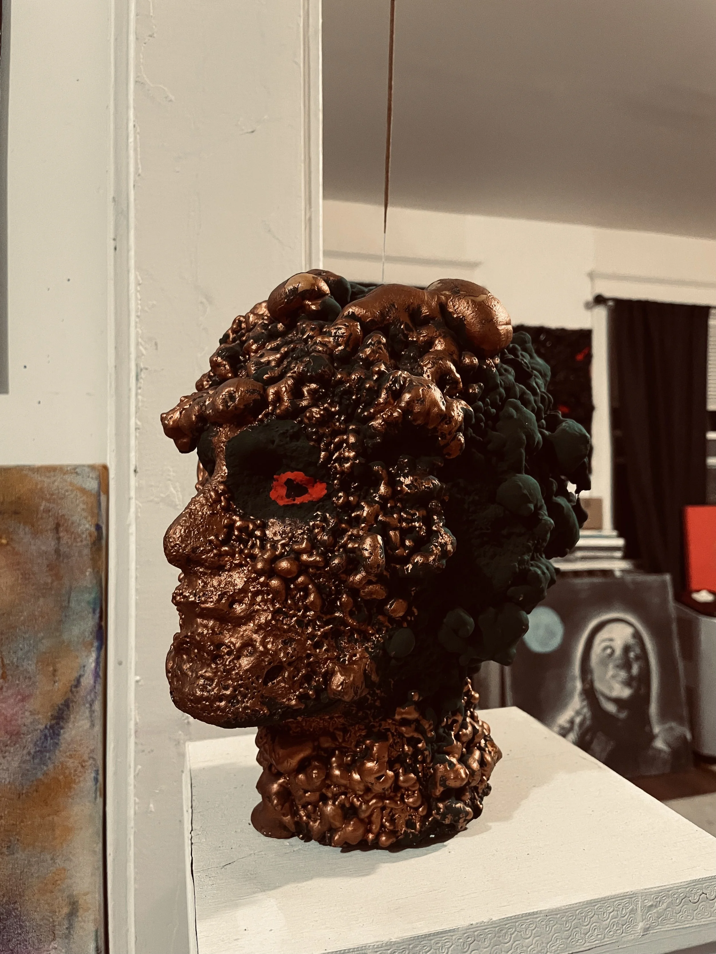 Sculpture of a human head made of various textured dark and metallic materials, displayed on a white pedestal in an art gallery.