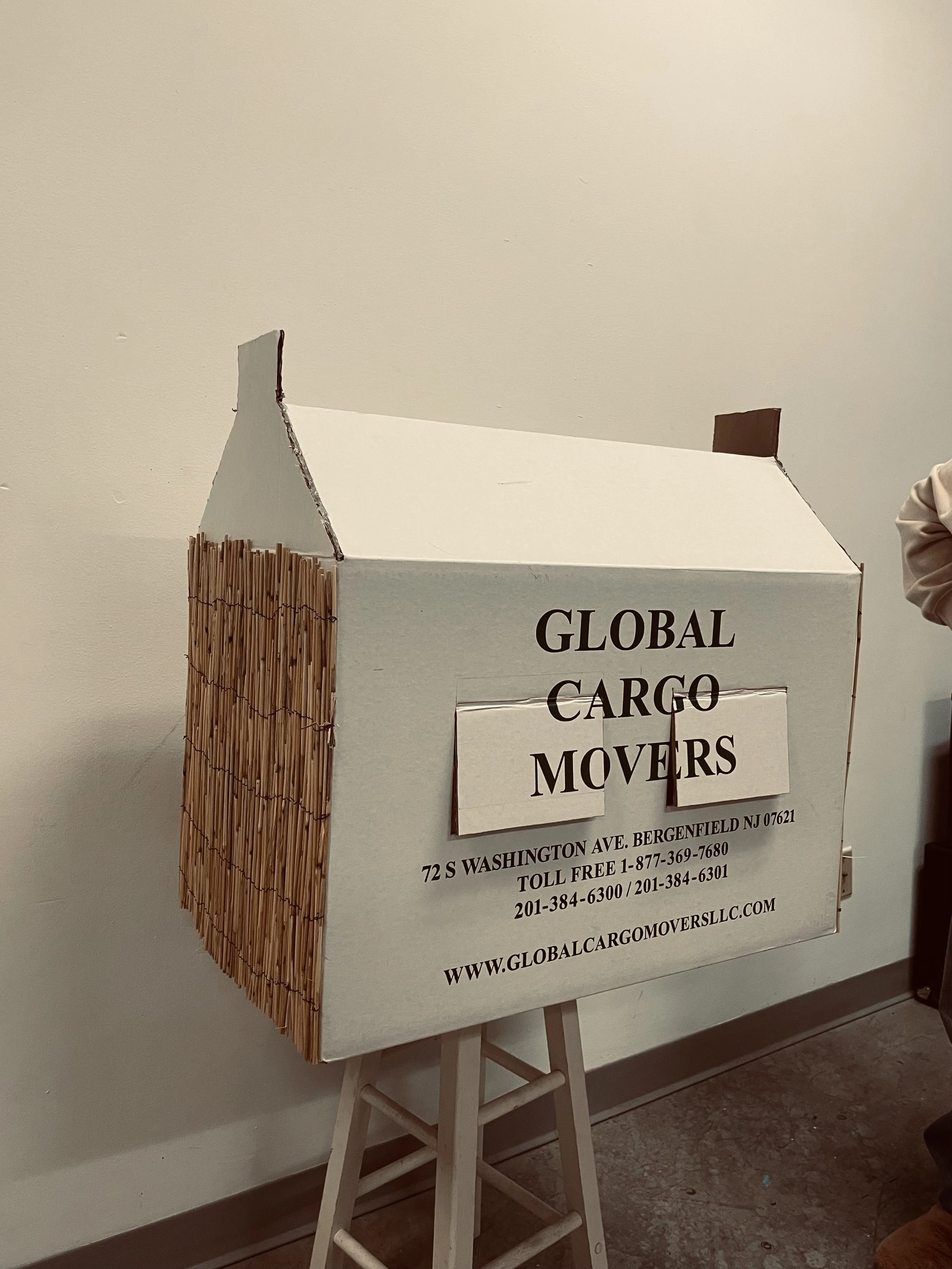 A cardboard standee shaped like a small house, advertising Global Cargo Movers with contact information and website, decorated with a bamboo mat on one side.