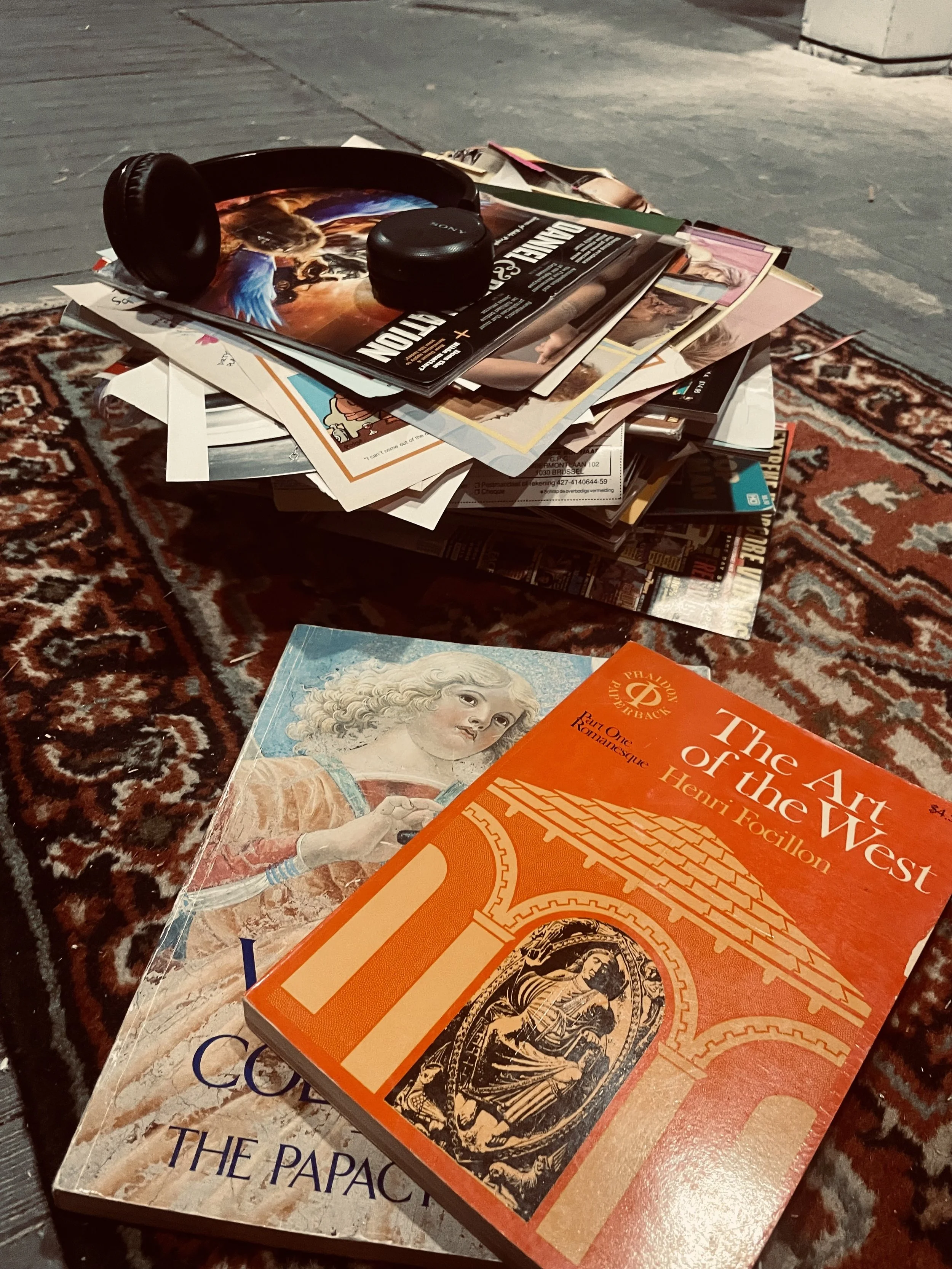 A pile of magazines and papers on an ornate, red patterned rug, with a pair of black headphones on top, in an indoor setting.
