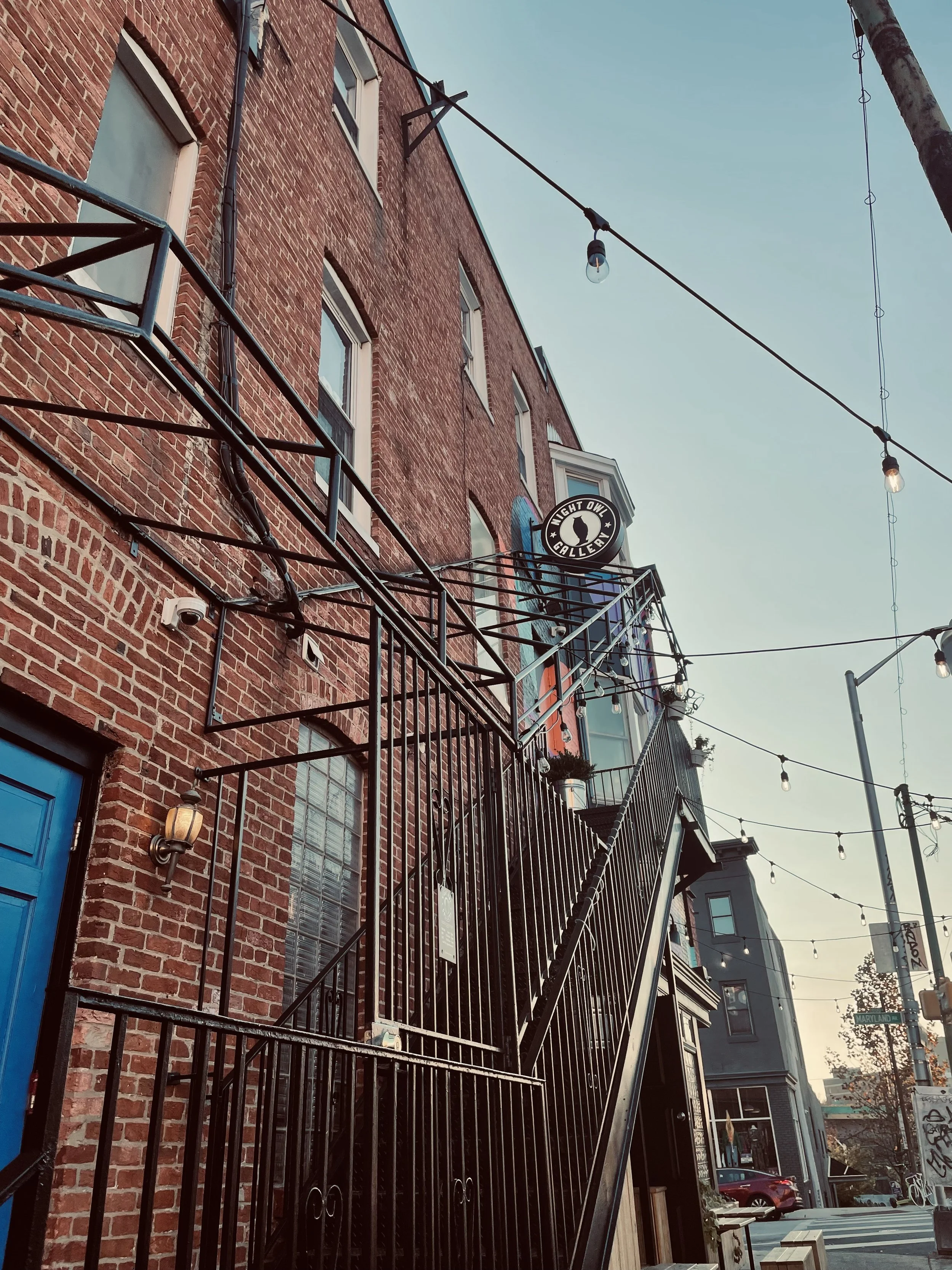 A brick building with an outdoor metal staircase leading up to a door. Strings of hanging lights are overhead, and there are signs and windows on the building. The sky is clear and blue.