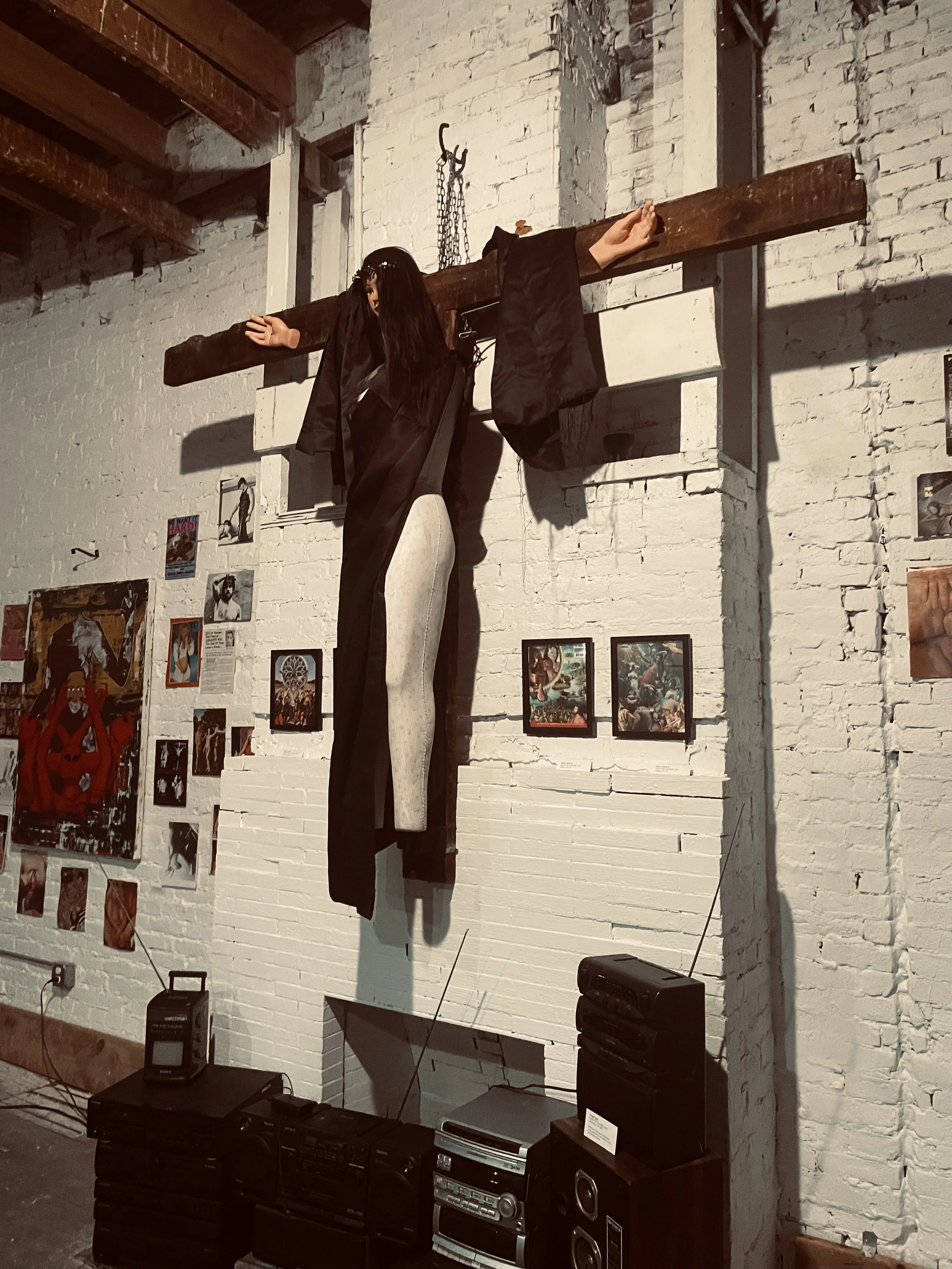 An art installation on a white brick wall depicting a suspended mannequin dressed as a monk with a wooden cross background, surrounded by smaller framed artworks, with a speaker and stereo system below.