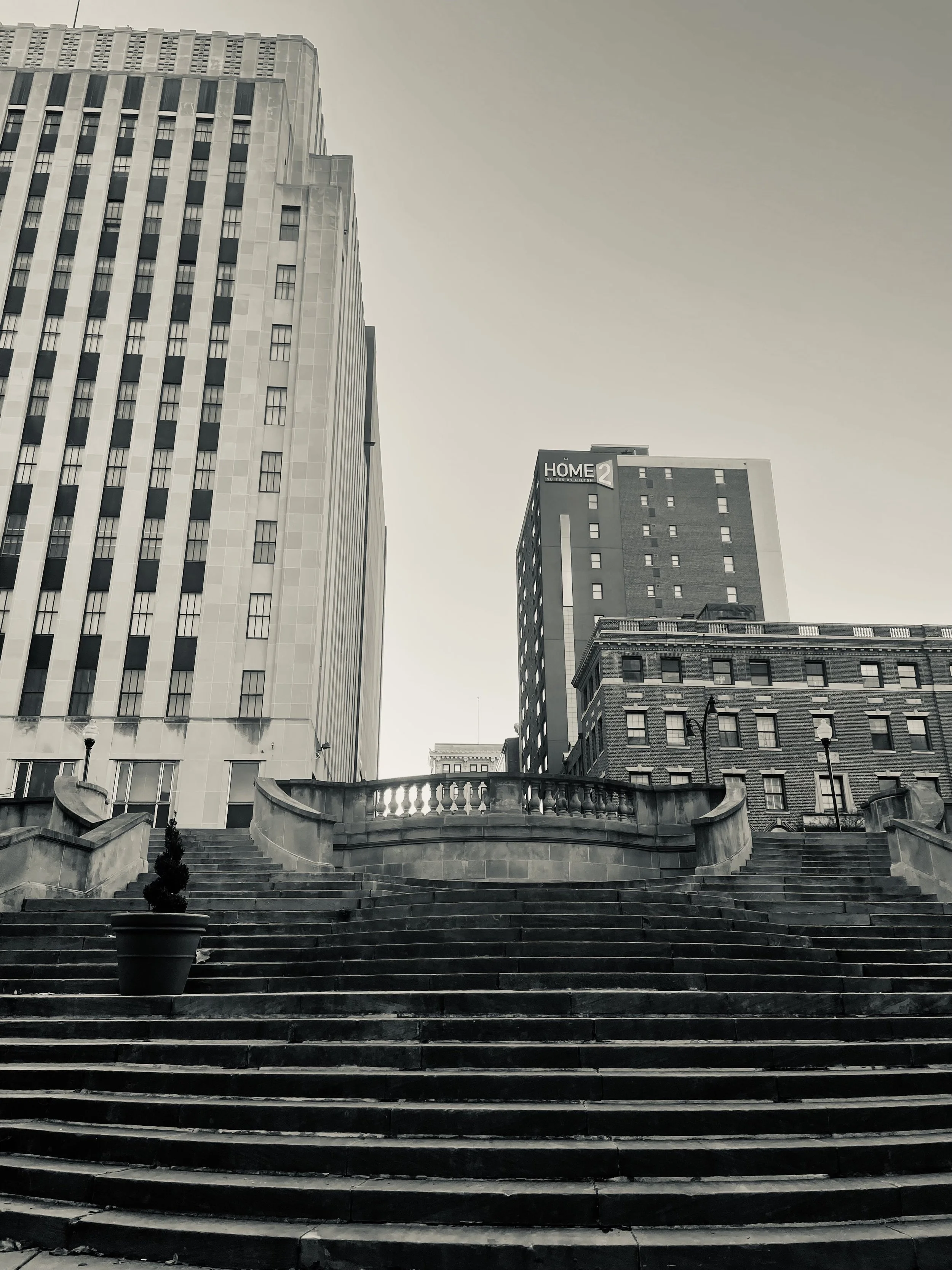 Cityscape with tall buildings, stone staircase, potted plant, and street lamps, in black and white.