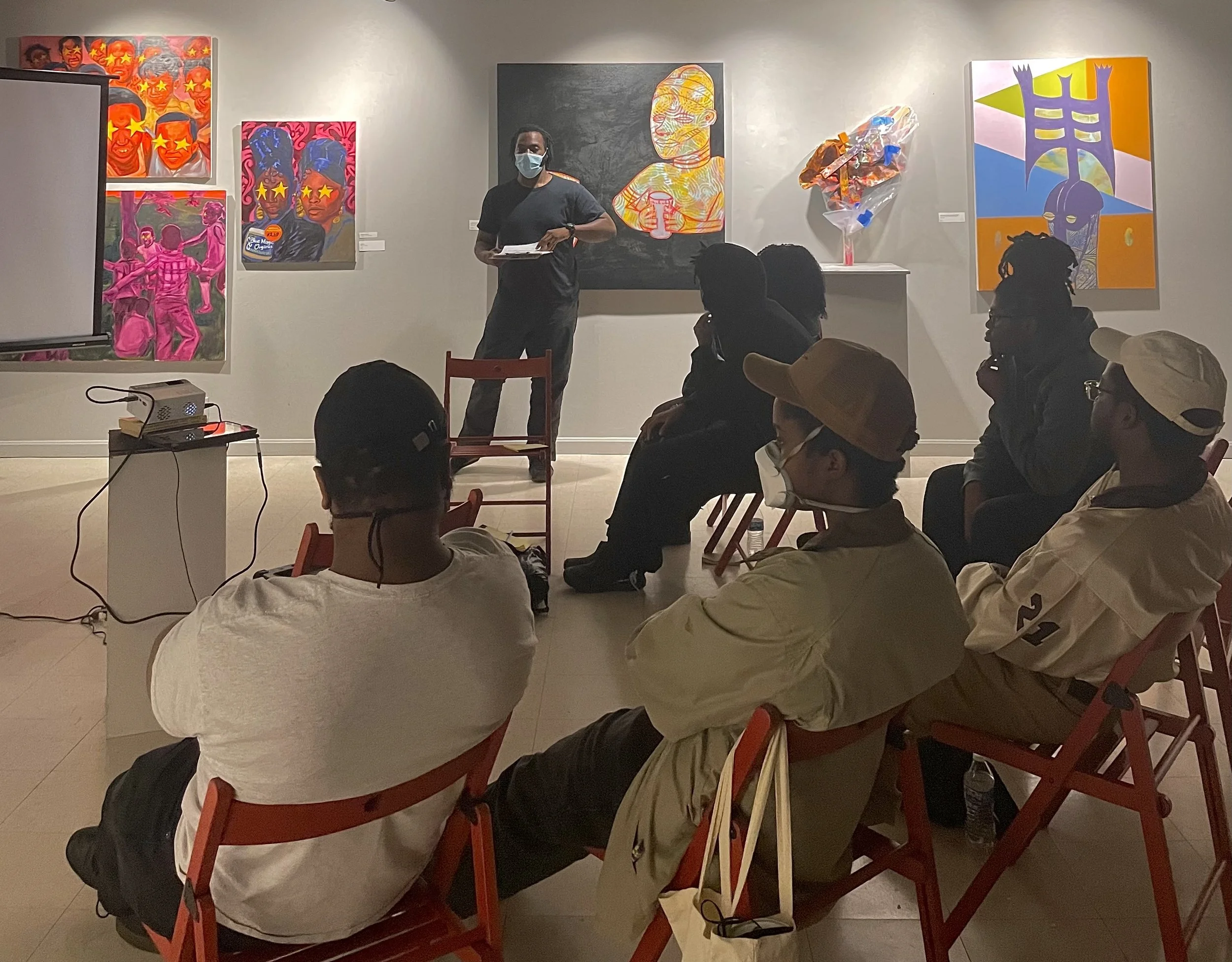 An art gallery with several colorful contemporary paintings on the wall. A woman wearing a mask is standing in front of the paintings and appears to be giving a presentation or lecture to a seated audience that includes men wearing masks and hats. Some audience members are attentively listening, while others are looking at the artwork.