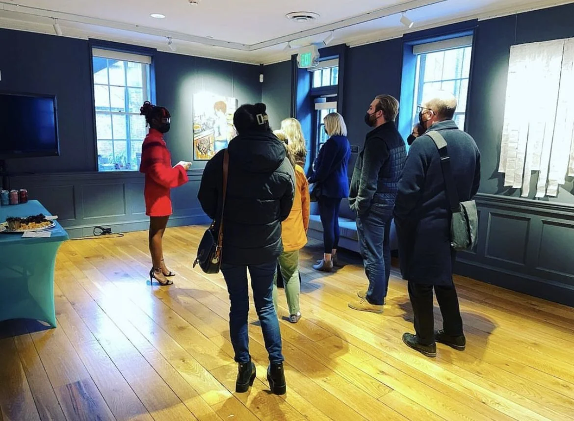 A group of people are standing in a line inside an art gallery, facing a woman in a red outfit who appears to be giving a presentation. The gallery has wooden floors, blue walls, large windows, and a painting on the wall behind the presenter.