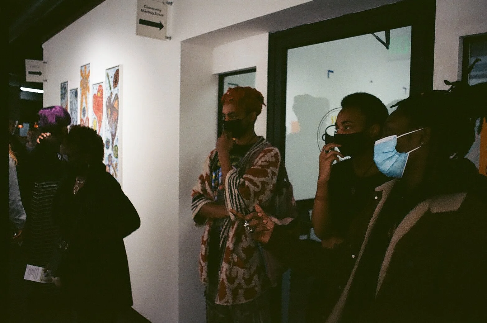 A group of people wearing face masks are standing in an art gallery, looking at colorful artwork on the wall.