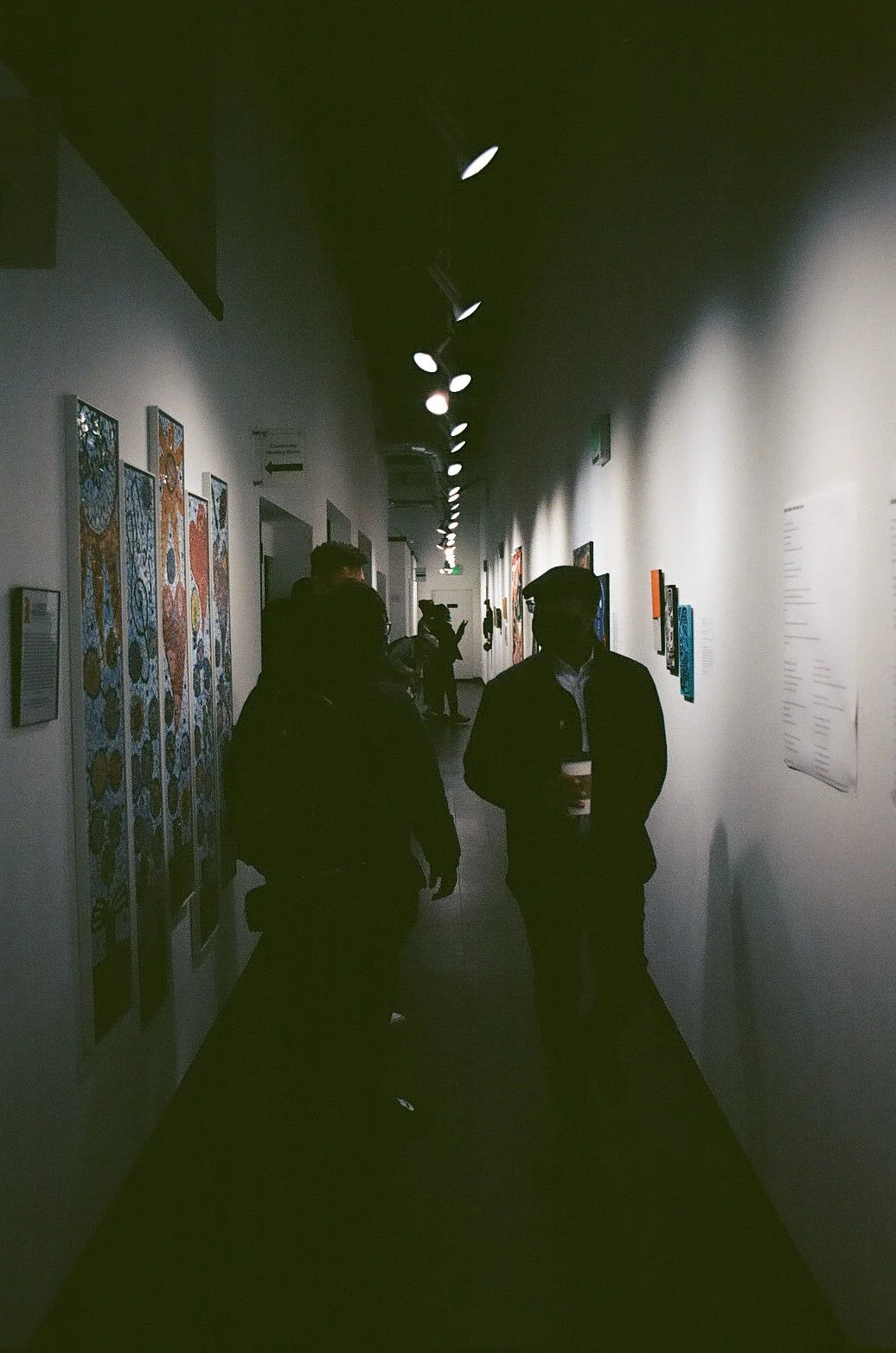 People walk through an art gallery at night, with artworks hanging on white walls and ceiling spotlights illuminating the space.