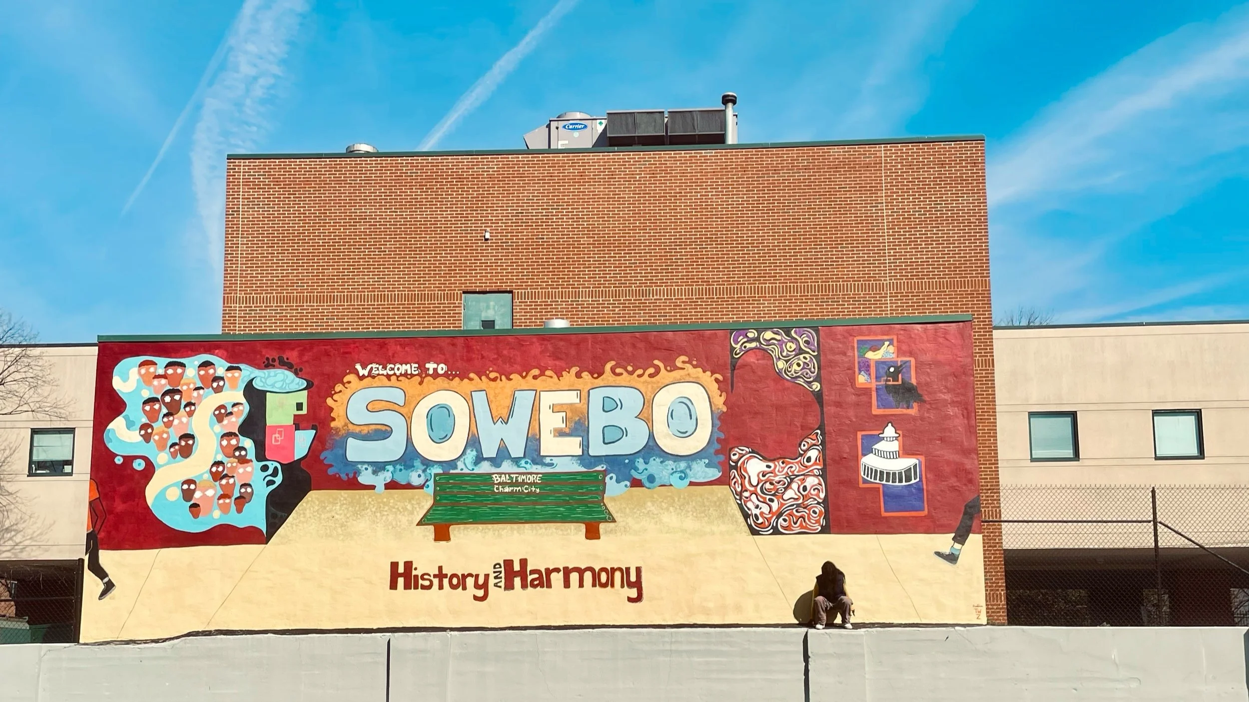 Colorful mural on building wall reading 'Welcome to Sowebo', featuring abstract art, a park bench with 'Baltimore Charm City' written on it, and the words 'History and Harmony' beneath.