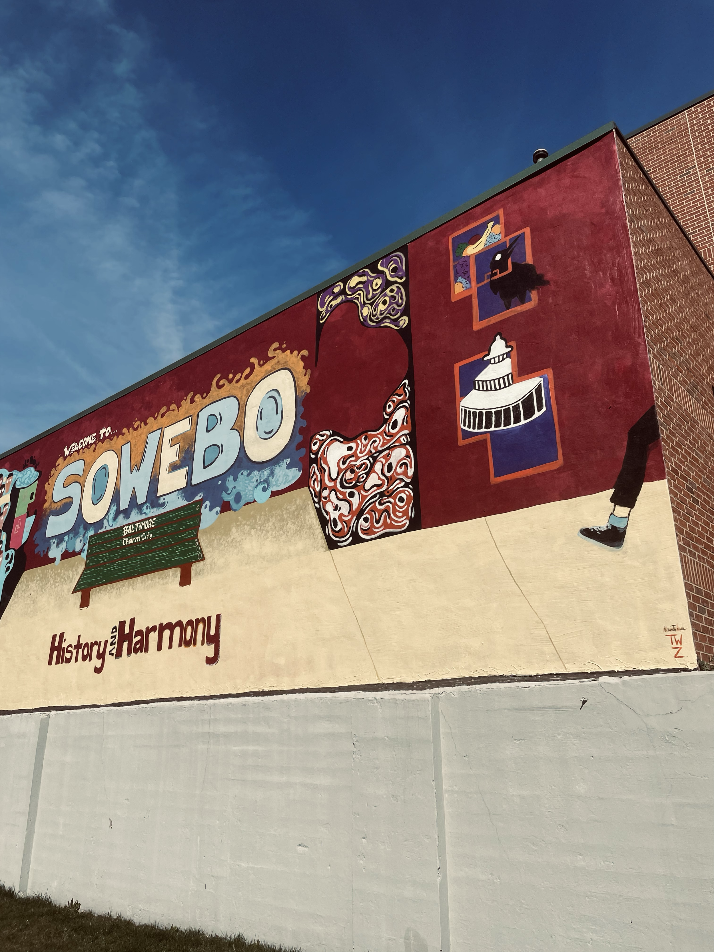 Colorful mural with the words 'Welcome to SOWEBO' and symbols representing Baltimore's Charm City. Includes artwork, city landmarks, and a person's leg and shoe on the right.