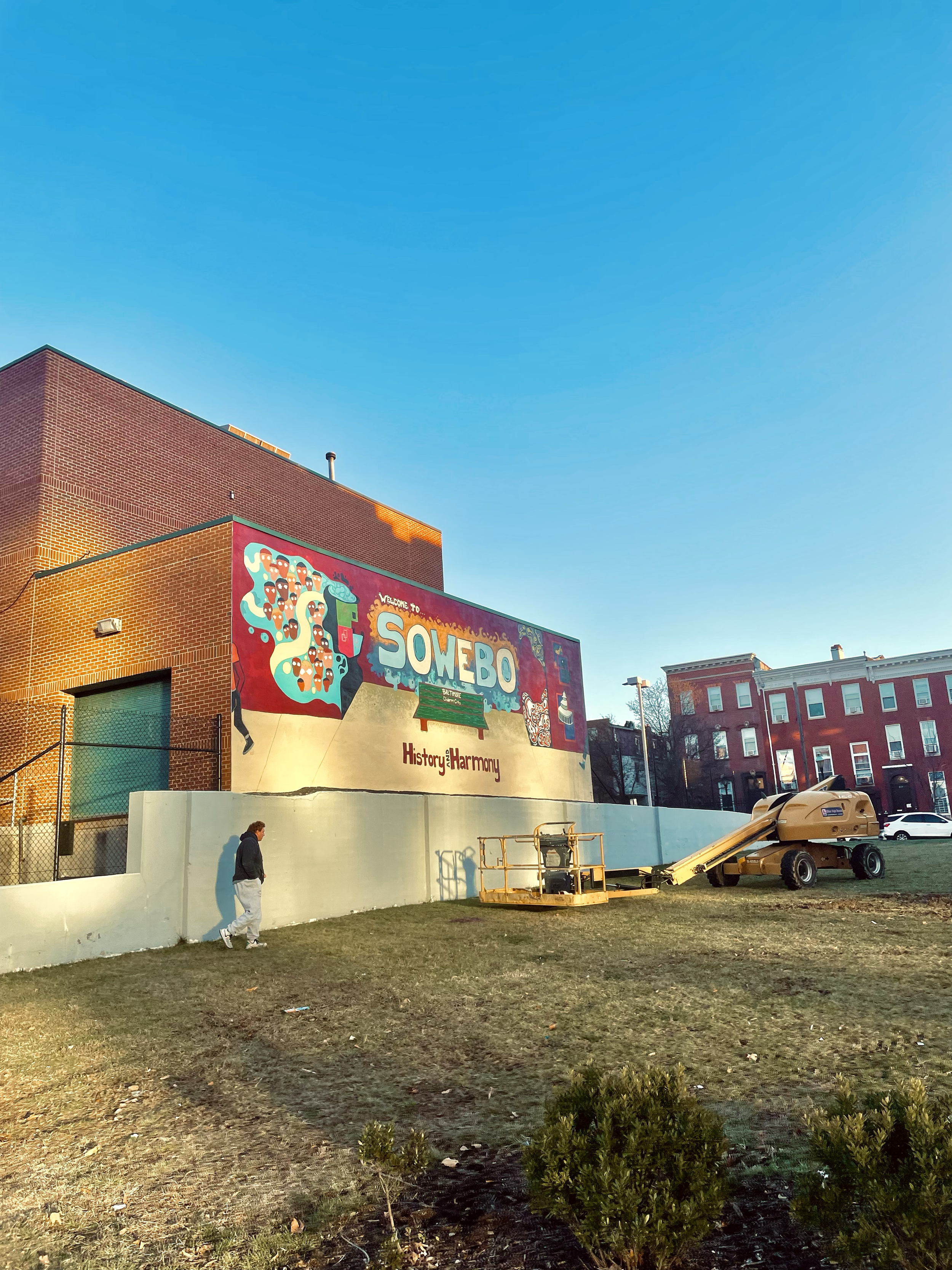 Colorful mural on a brick building welcoming visitors to SoweBo, with the words 'History & Harmony.' In front, there is a man walking and construction equipment on the grass.