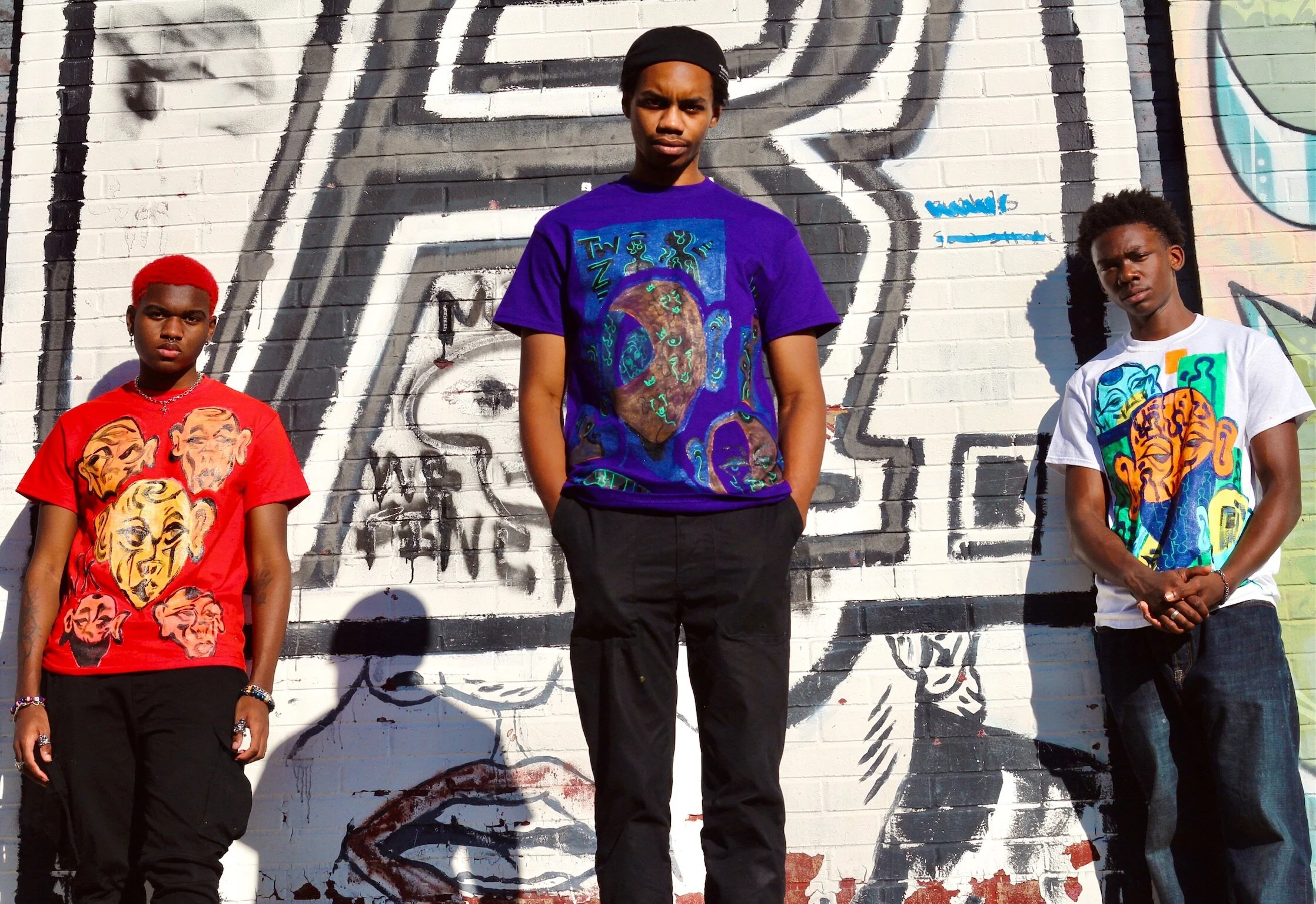 Three young men stand in front of a graffiti wall. They are wearing colorful graphic t-shirts, with the middle one in a purple shirt, the left one in red, and the right one in white. The graffiti features stylized faces and abstract designs.