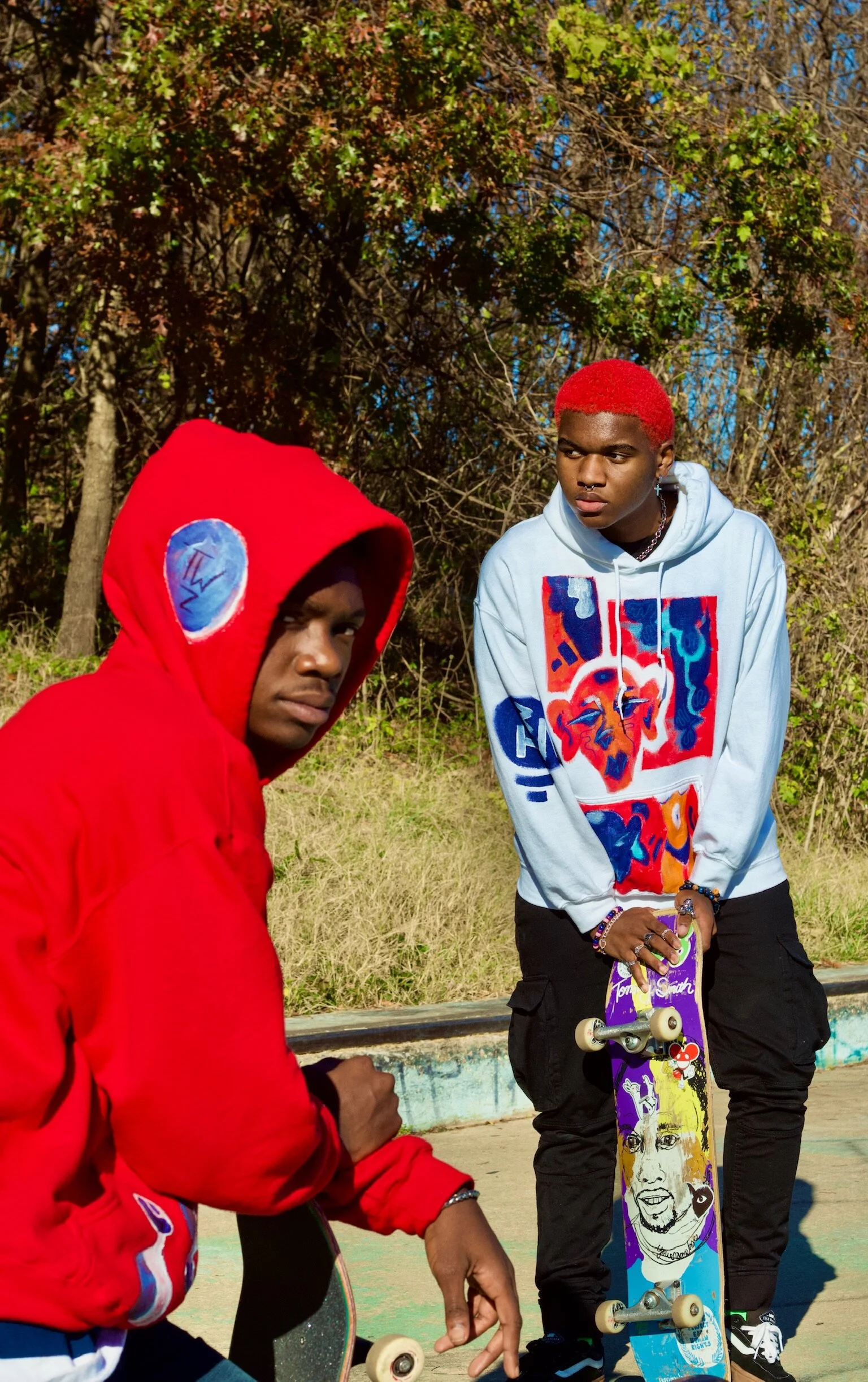 Two young men with skateboards outdoors; one wears a red hoodie with patches, the other a colorful graphic hoodie with red dyed hair, standing on a skate ramp with trees in the background.
