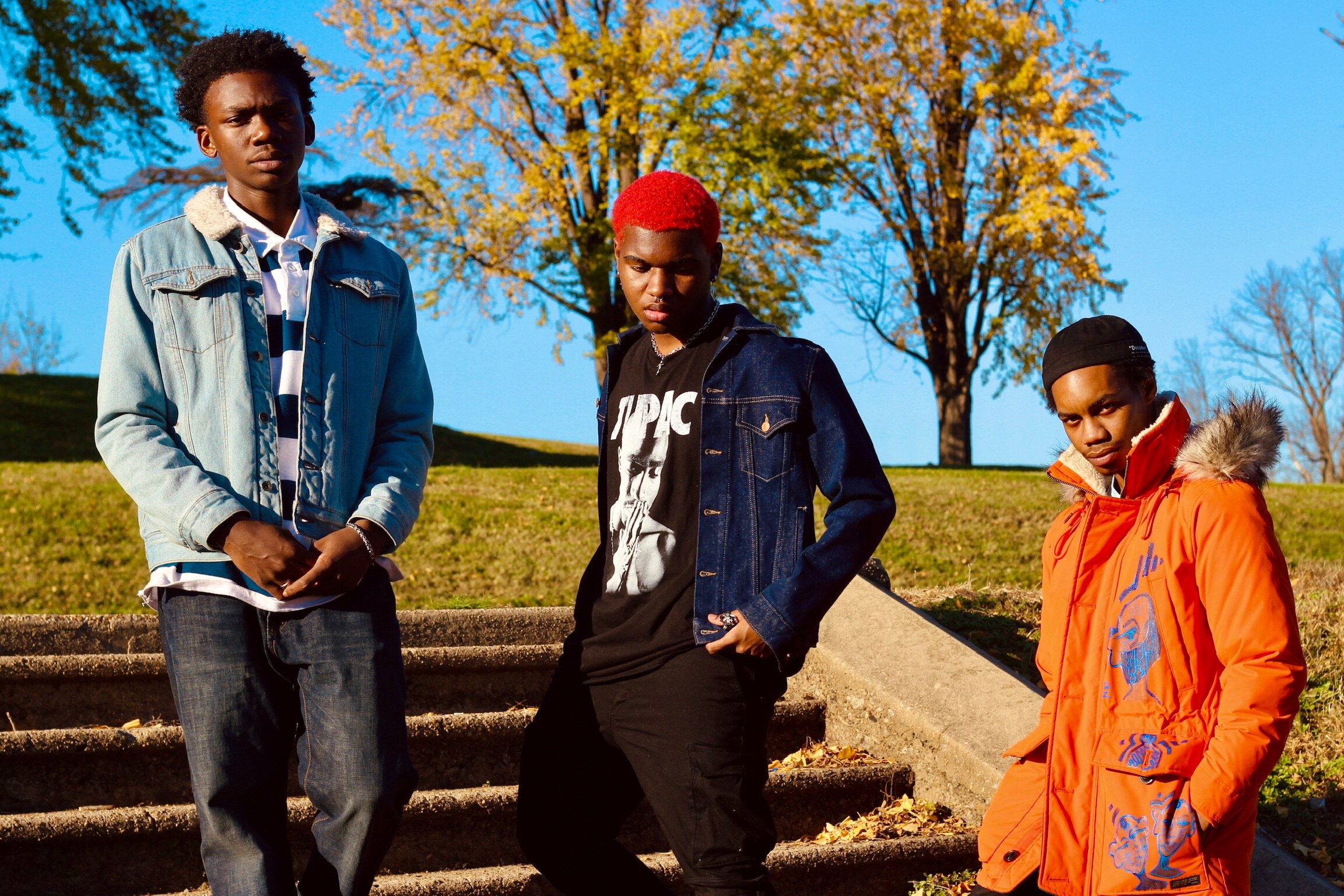 Three young men standing outdoors on steps with autumn trees and blue sky in the background.