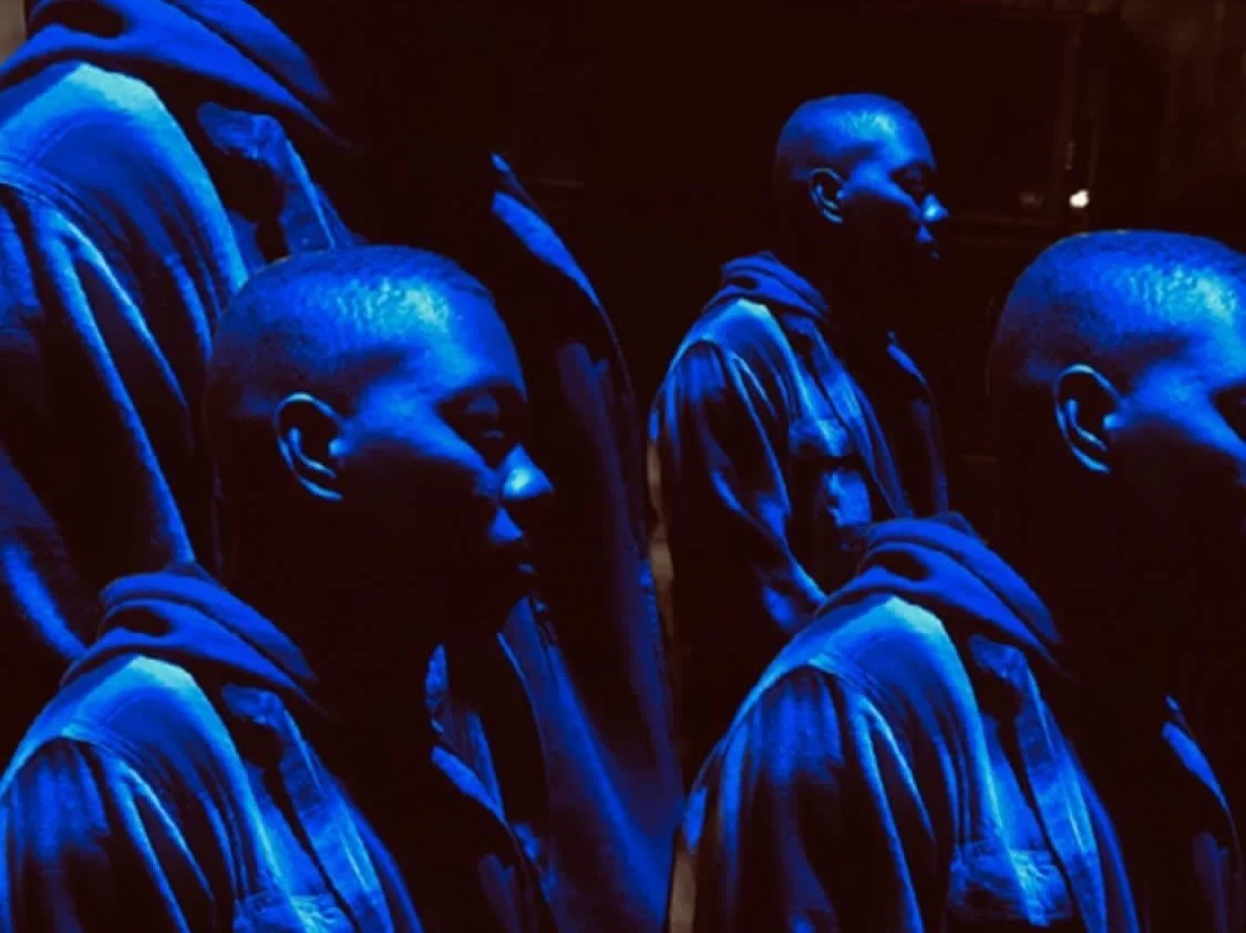 Multiple young men with short haircuts, dressed in hoodies, standing in dim blue lighting, appearing to be in a contemplative or serious mood.