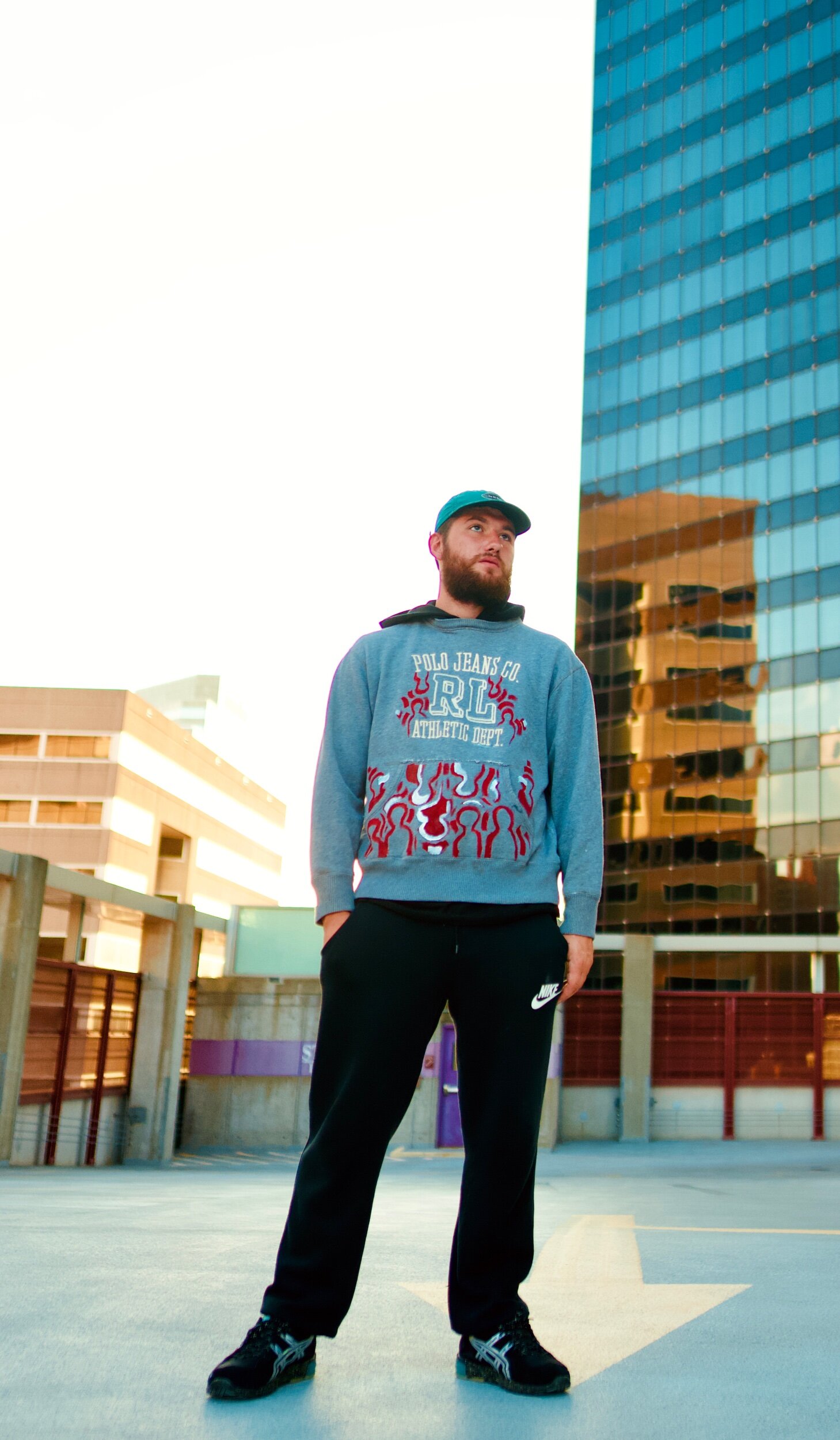 A man with a beard and blue cap standing on a rooftop parking lot, wearing a blue hoodie with red and white graphic design and black Nike sweatpants, with tall modern buildings in the background.