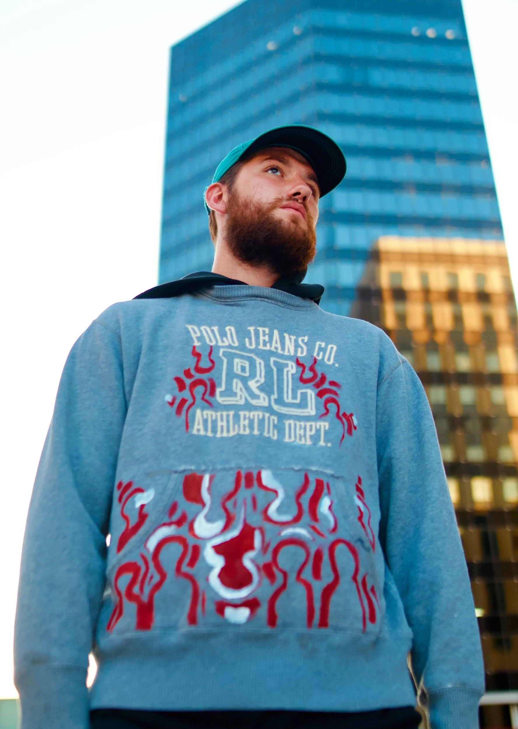 A young man with a beard and a teal cap looking upwards, standing outdoors in front of a tall modern glass building during daytime.