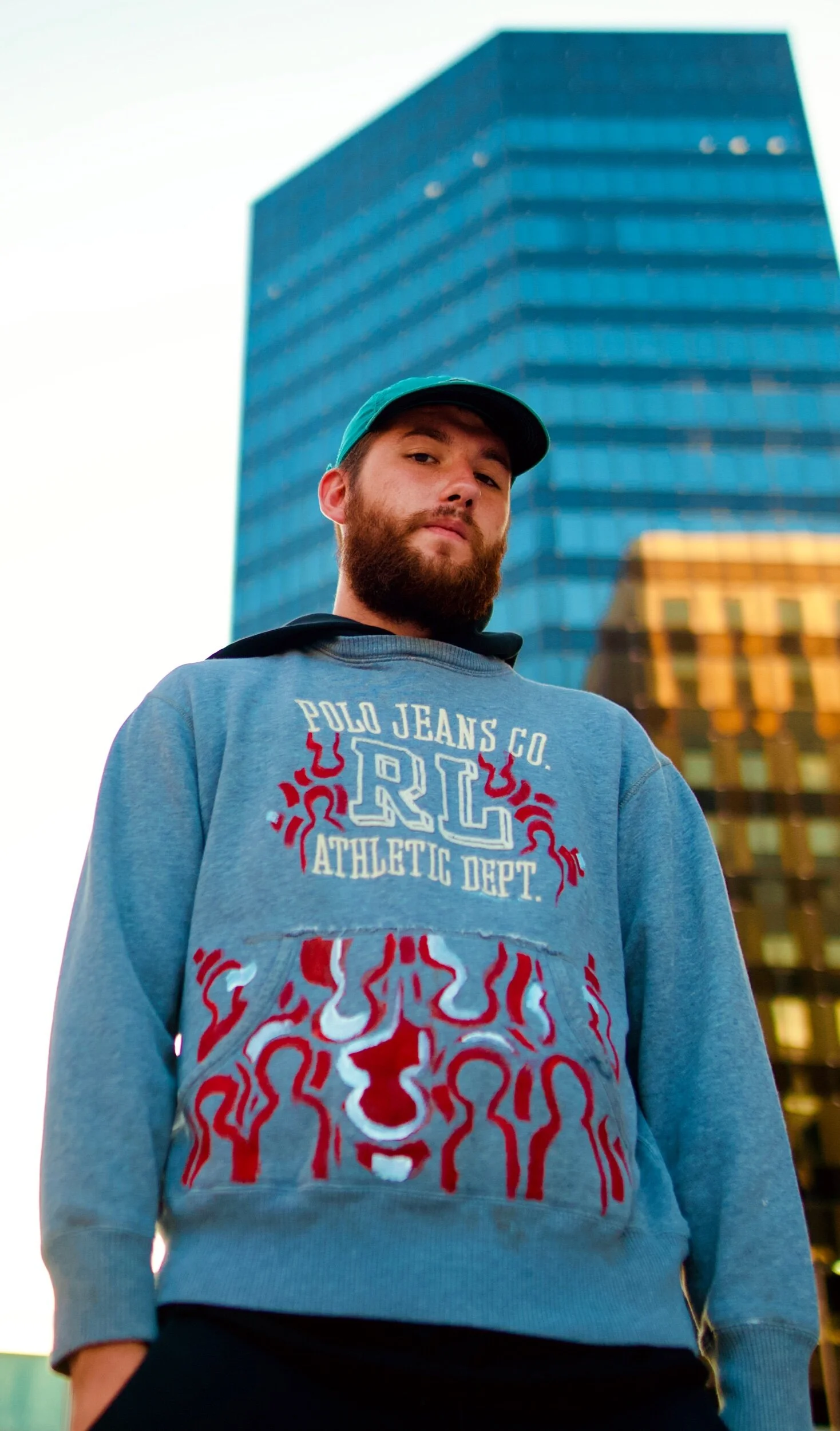 A man with a beard and baseball cap standing outdoors in front of a tall modern glass building.