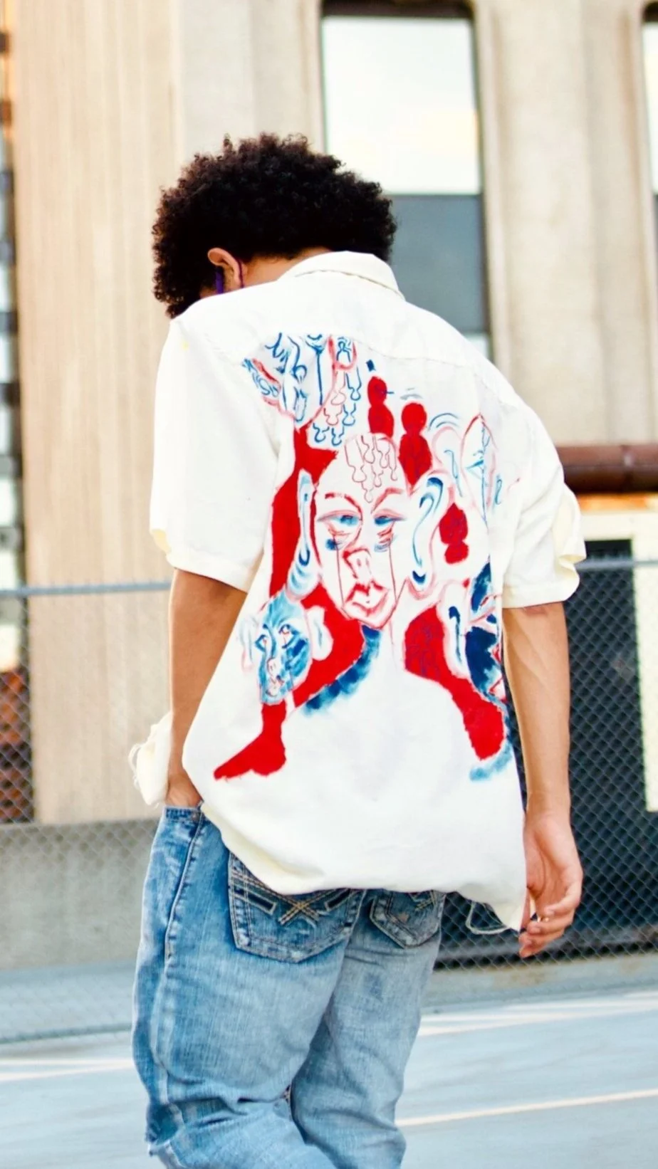 Person with curly hair wearing a white shirt with colorful abstract artwork on the back, standing outdoors near a chain-link fence.