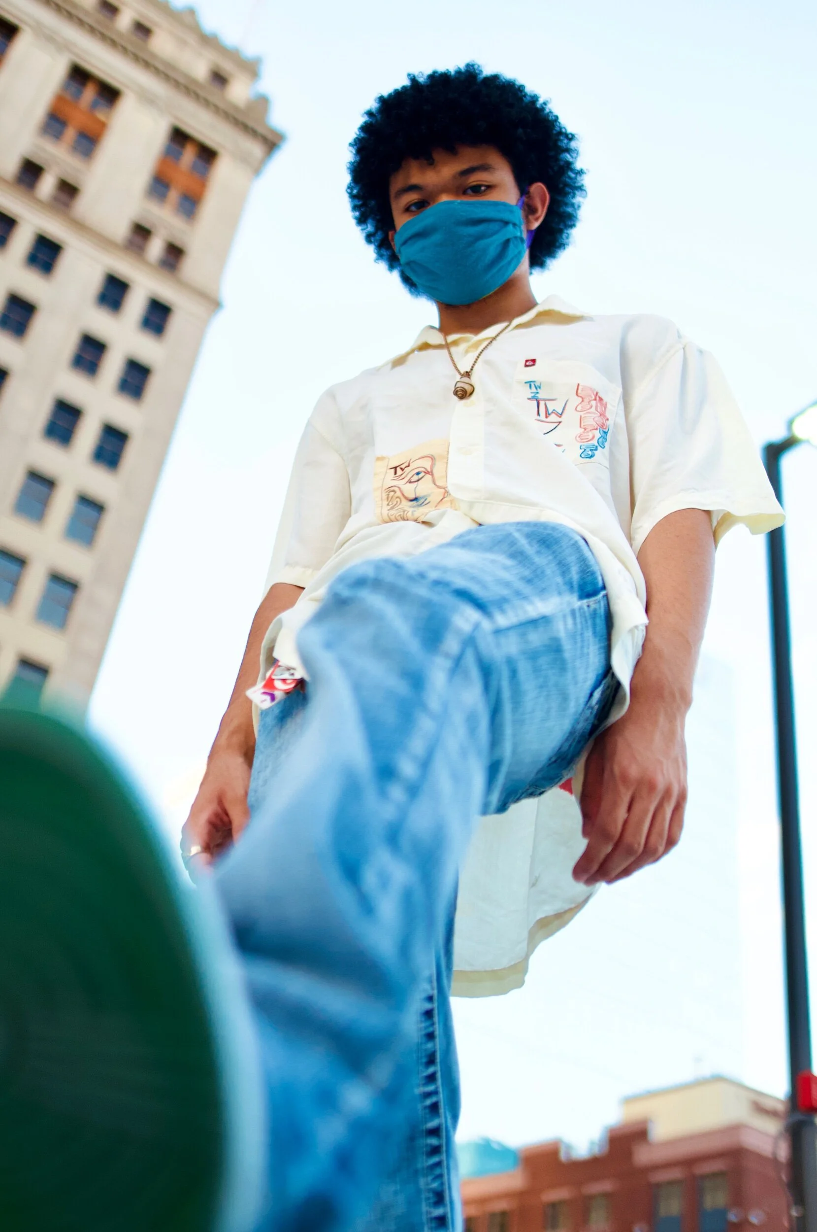 Young man wearing a blue face mask, white graphic shirt, and jeans, standing outdoors in an urban area during daytime, with a tall building and street lamp in the background.