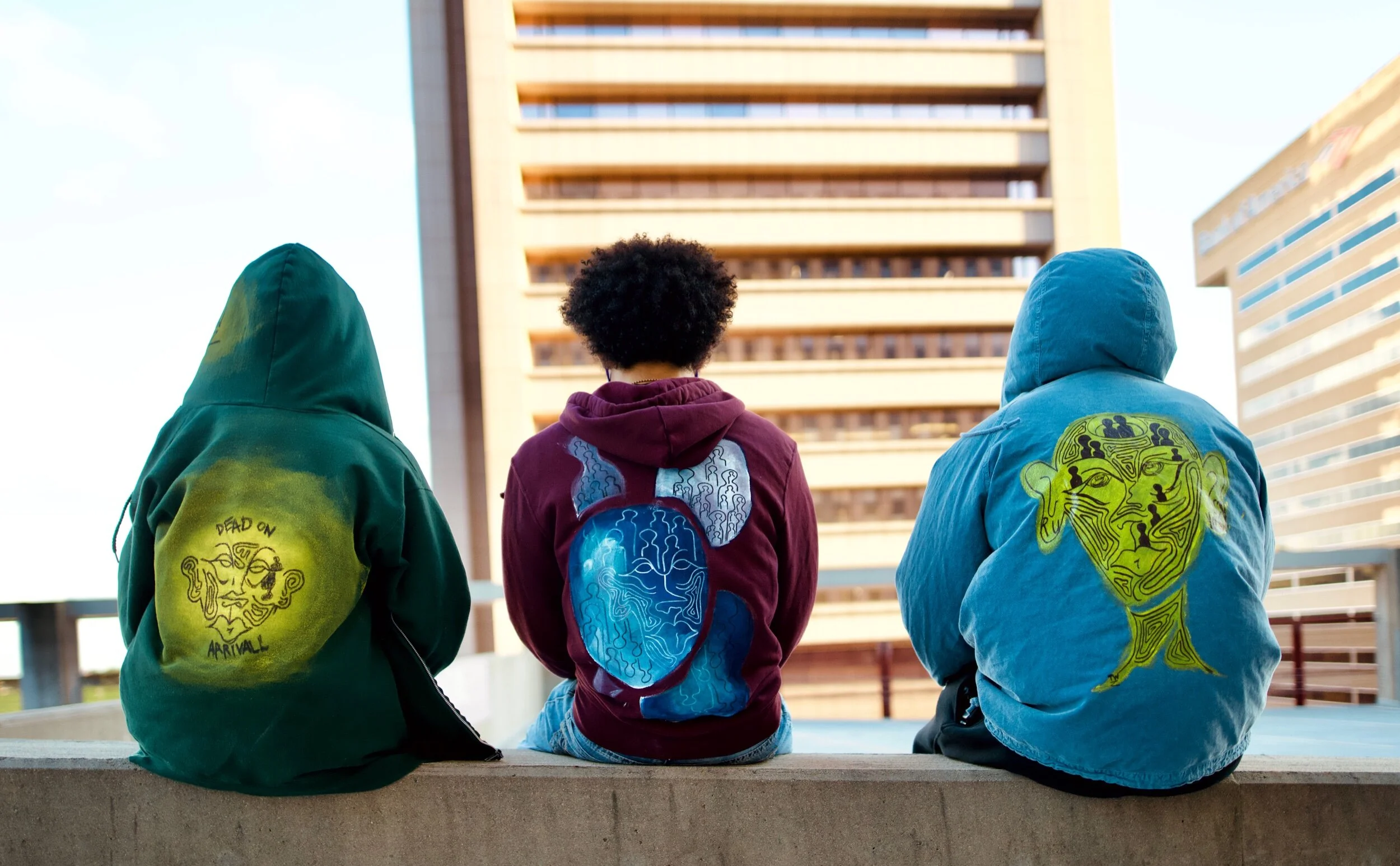 Three people sitting on a concrete ledge with their backs facing the camera, two wearing hooded jackets with artistic designs on the back and one with a backpack, in an urban setting with tall buildings.