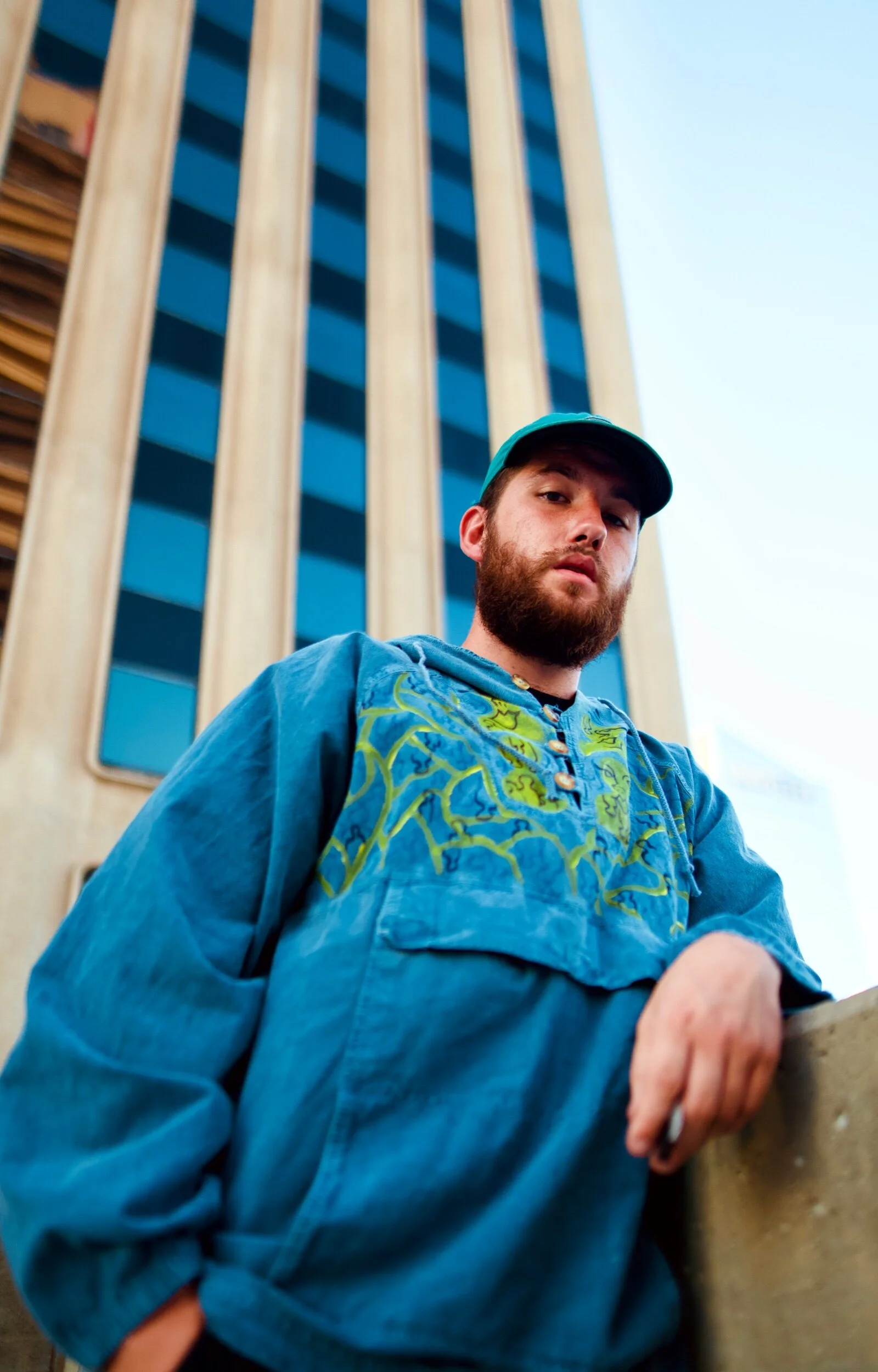A young man with a beard wearing a blue hoodie and cap, leaning on a wall with a tall building in the background.