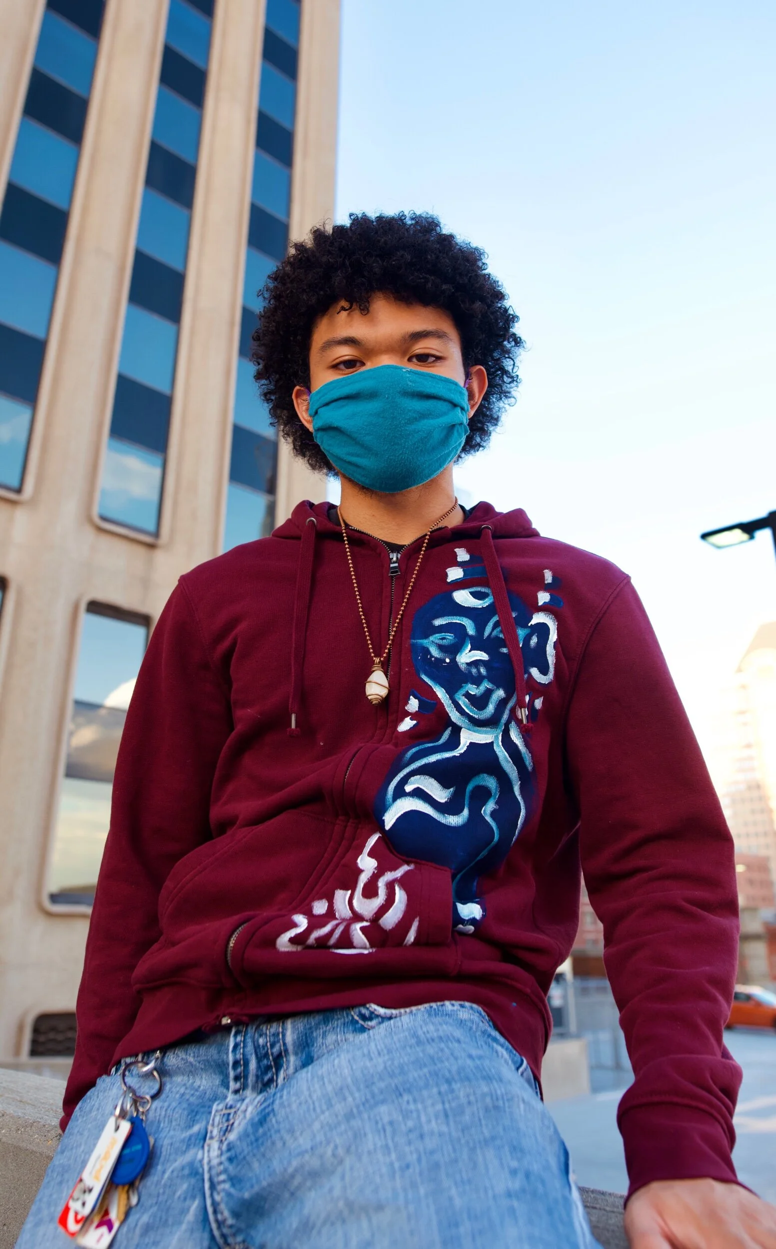 A person wearing a burgundy hoodie with a blue and white abstract face design, blue jeans, and a blue face mask sitting outdoors in an urban area with buildings and a blue sky in the background.
