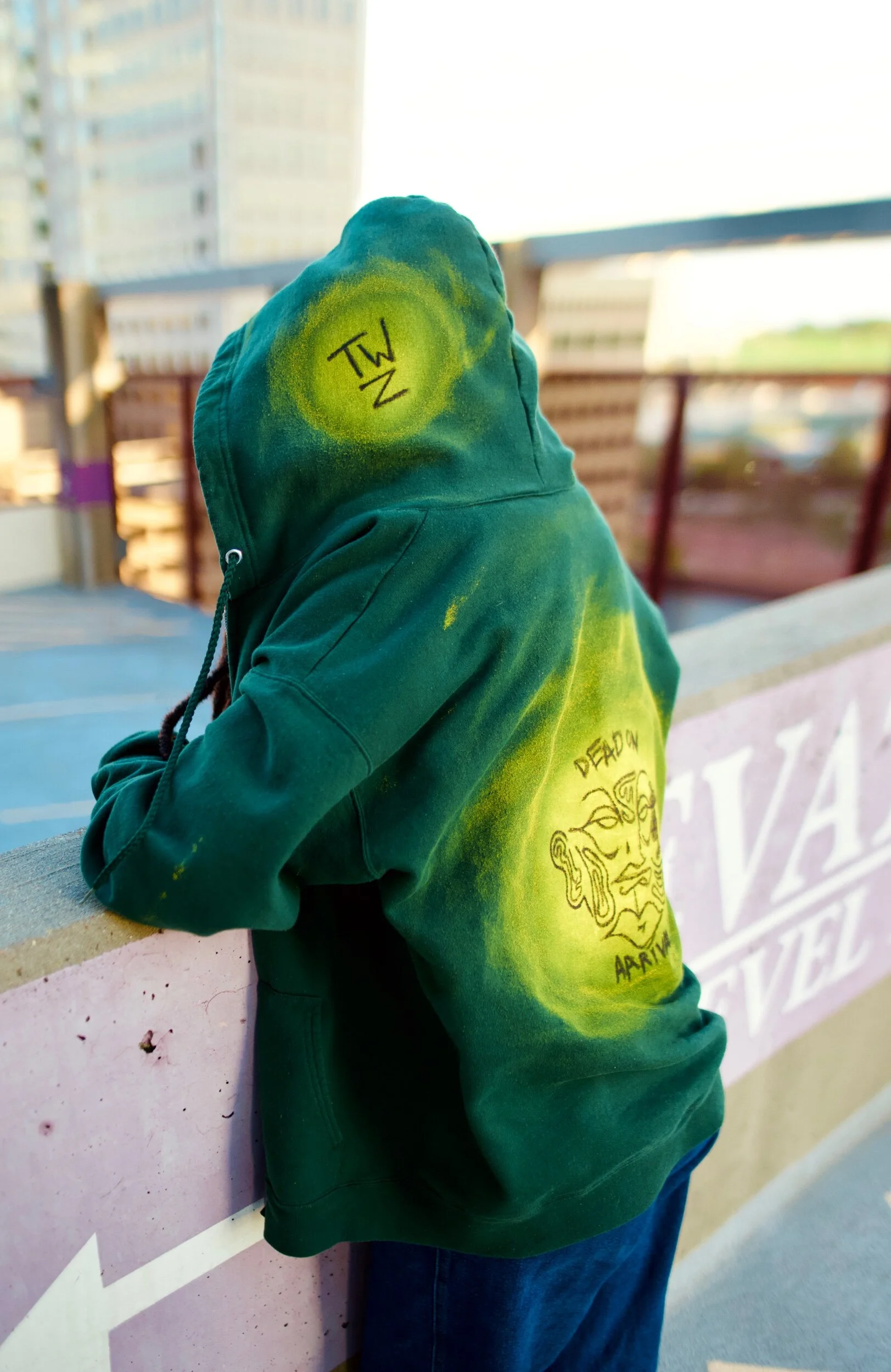 Person wearing a green hoodie with yellow spray paint accents and a drawing of a face on the back, leaning on a concrete barrier in an outdoor urban setting.