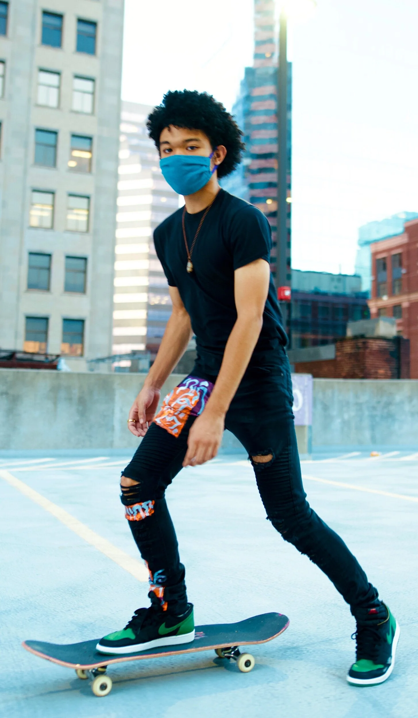 A young man with afro hair skateboarding on an outdoor parking garage with tall city buildings in the background. He is wearing a blue face mask, black t-shirt, ripped black jeans, and green Nike sneakers.