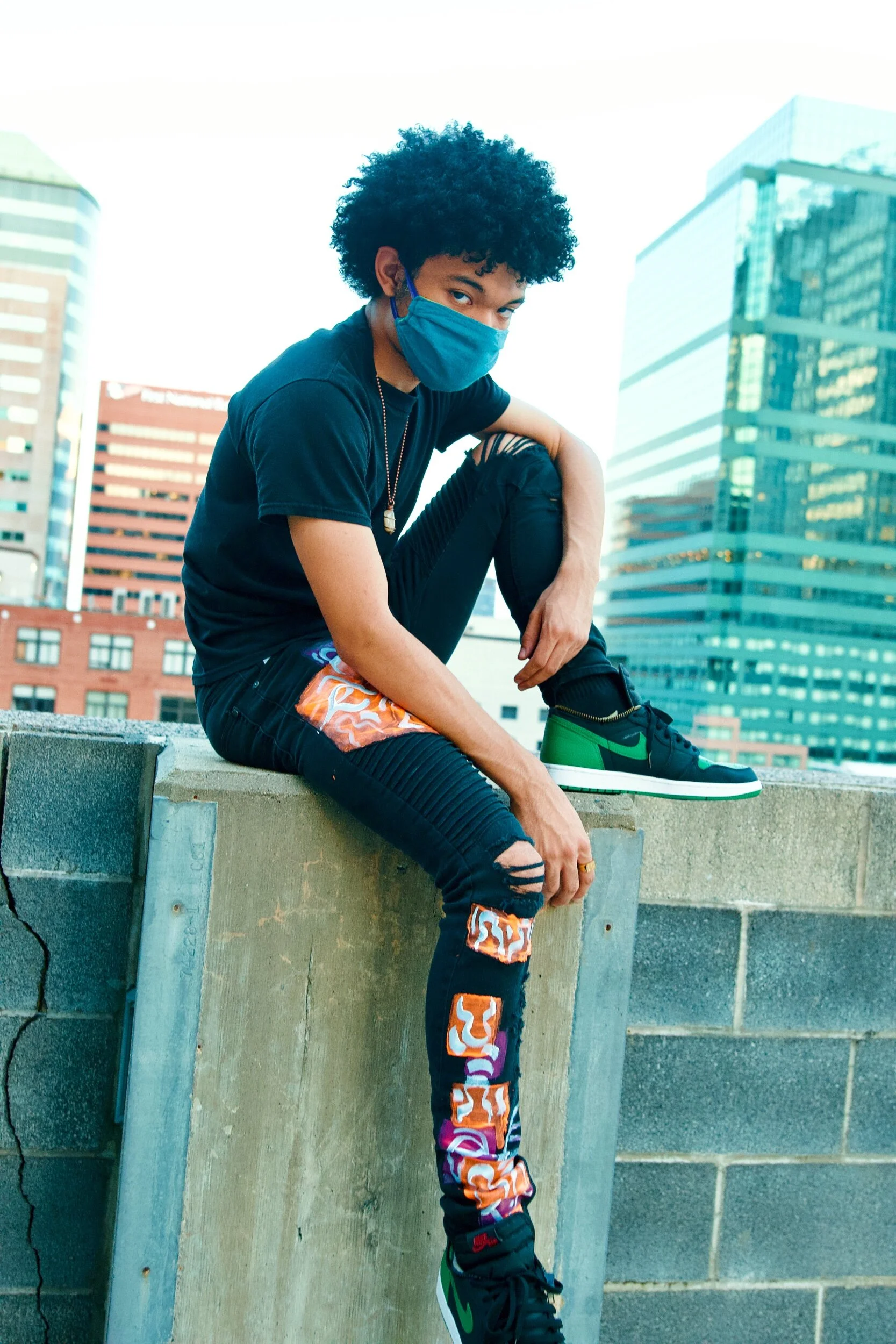 A young man with curly hair wearing a blue face mask, black t-shirt, ripped black jeans with colorful patterns, and green and black sneakers is sitting on a rooftop ledge in an urban area with tall buildings in the background.