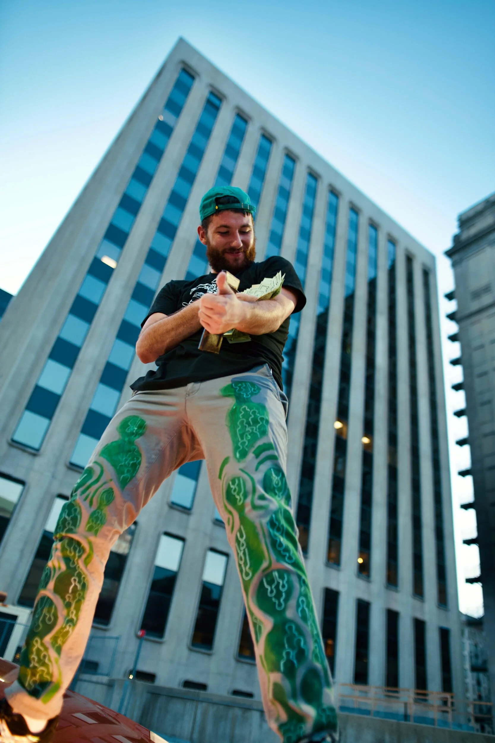 A man with a beard wearing a green cap, black t-shirt, and colorful painted pants is standing outdoors in front of a tall building, holding a stack of money and giving a thumbs-up.