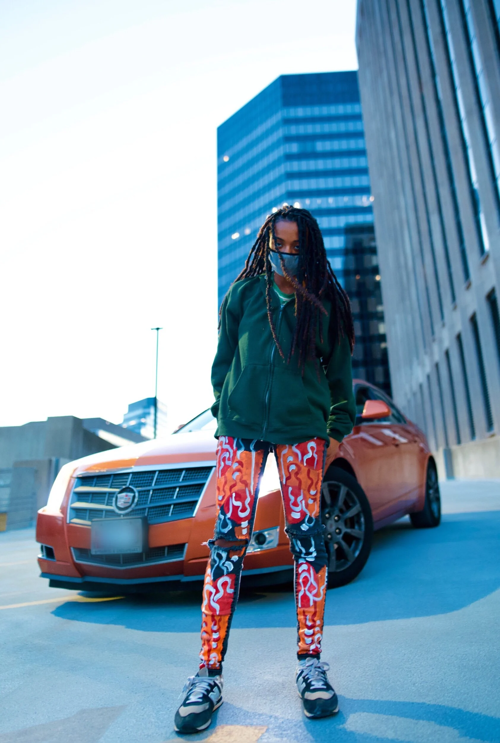 A woman with dreadlocks wearing a face mask, green jacket, vibrant patterned pants, and sneakers standing in front of an orange car on a city street with tall office buildings.
